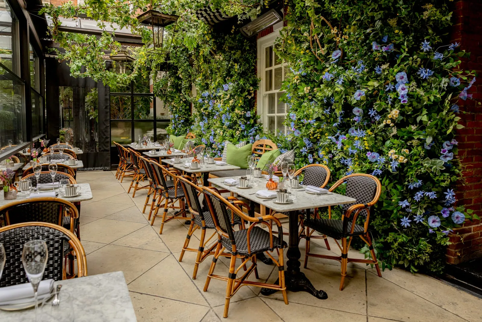The same terrace in summer, green climbing plants and blue flowers on the walls over the same marble tables and rattan chairs