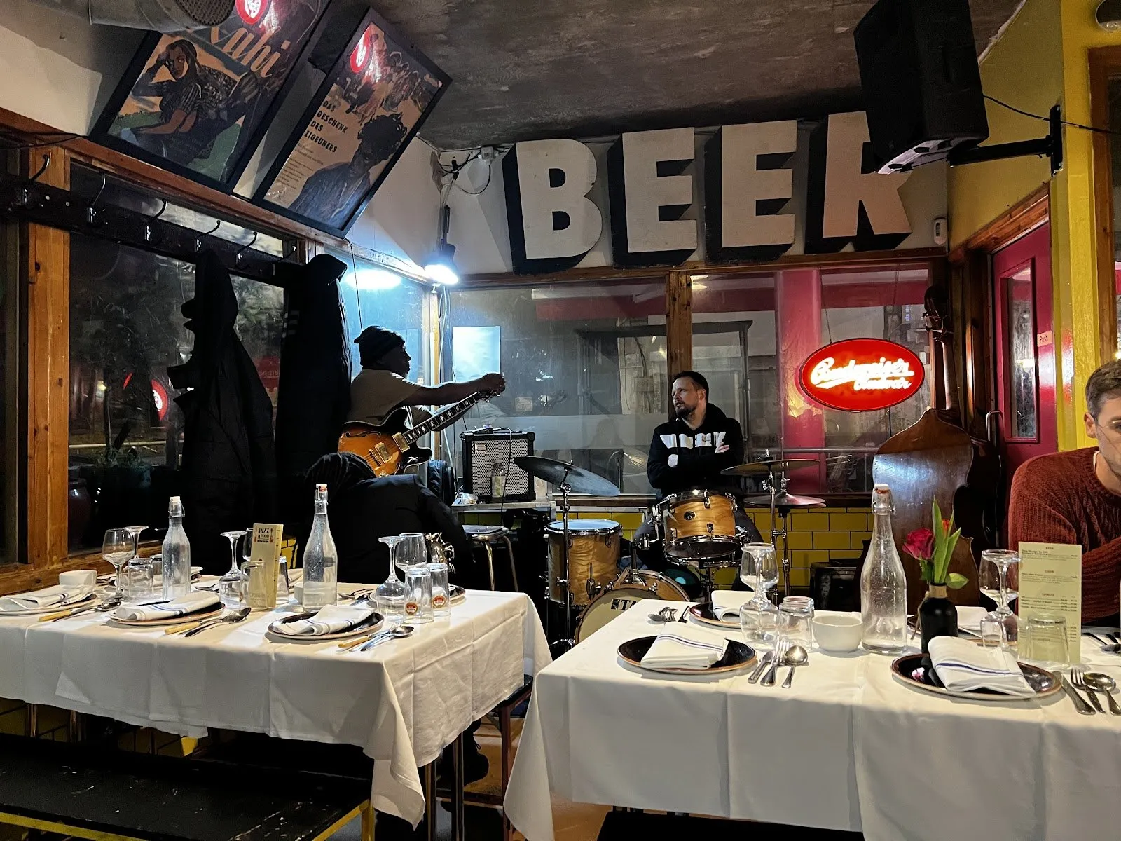 A guitarist and a drummer play among tables laid with white cloths at Dalston Jazz Bar, under a large BEER sign