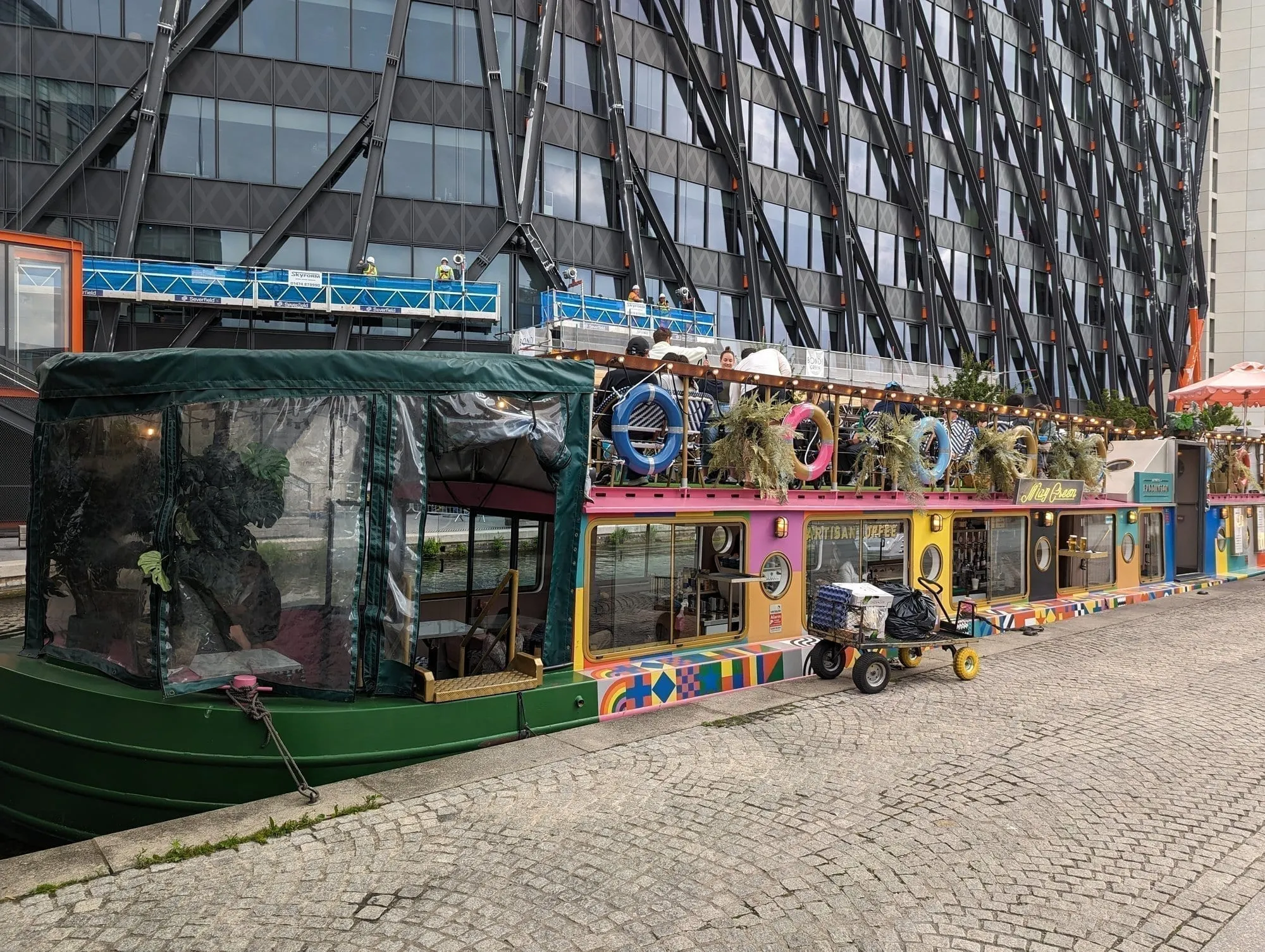 The brightly painted narrowboats of Darcie and May Green moored at Paddington Basin, with a rooftop terrace of diners above