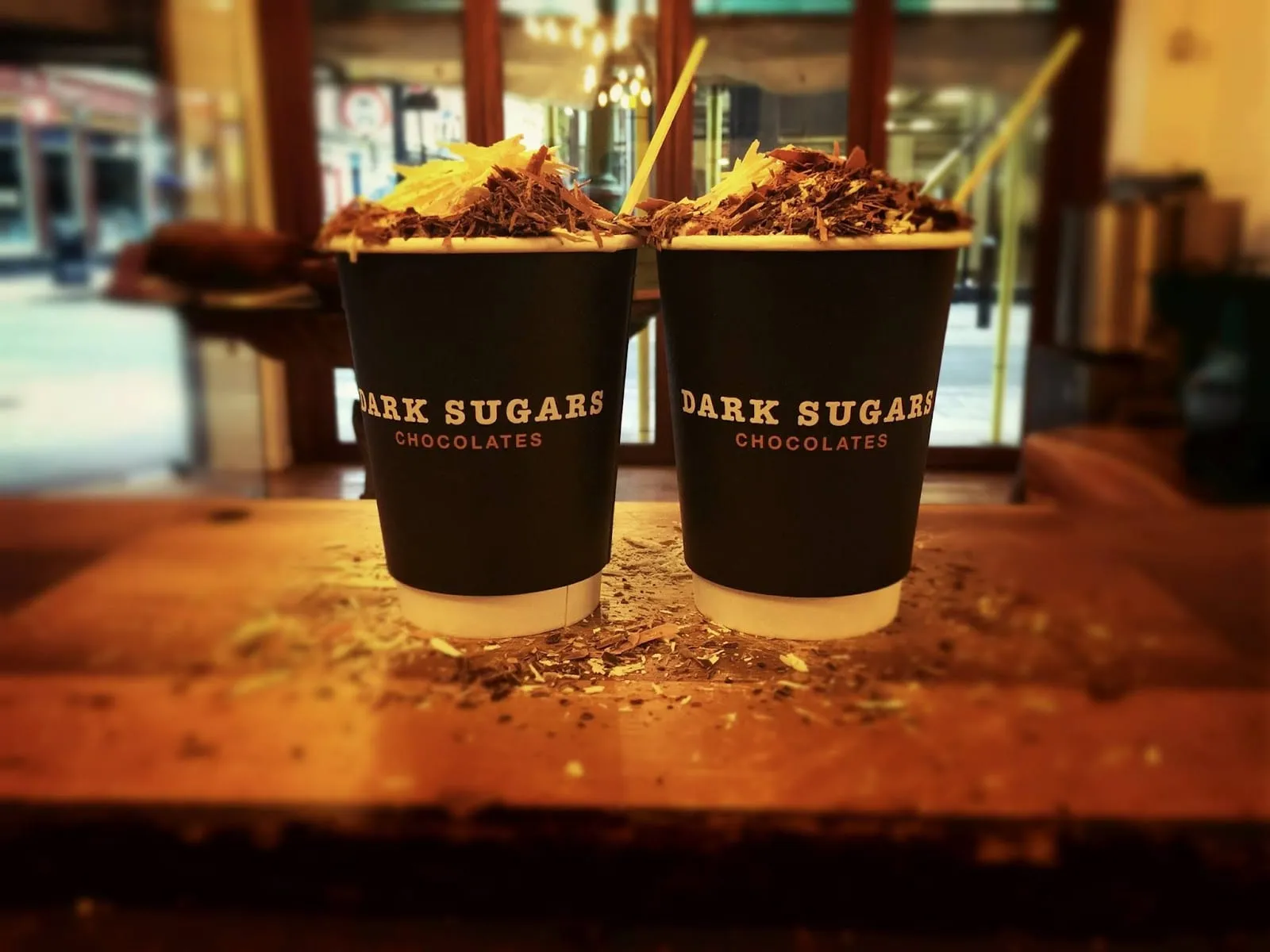 Two takeaway cups from Dark Sugars Cocoa House piled high with chocolate shavings, standing on a wooden counter scattered with fallen curls of chocolate