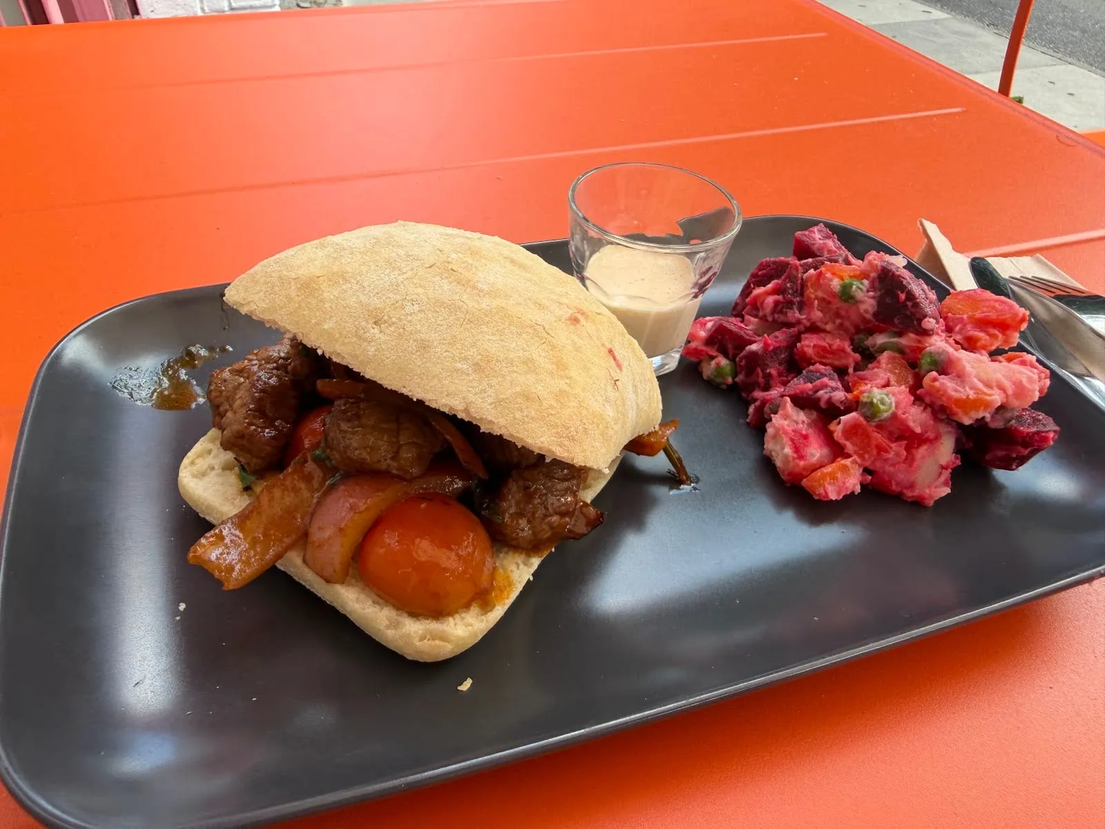 A crusty white roll filled with stir-fried beef, red onion and tomato on a black plate, beside a shot glass of pale sauce and a pile of diced beetroot, carrot, potato and peas in mayonnaise