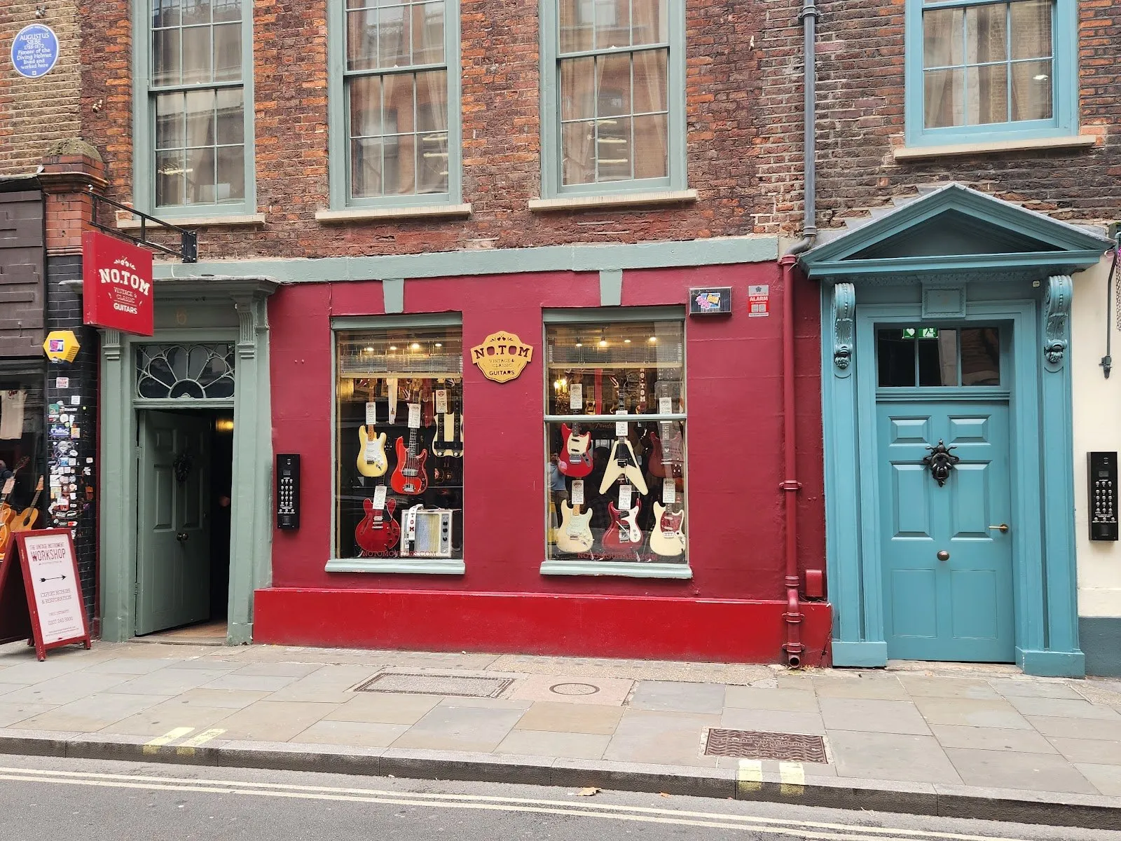 The red shopfront of No.Tom vintage guitars at 6 Denmark Street, electric guitars hanging in both windows, with the blue Augustus Siebe plaque high on the brick building to its left