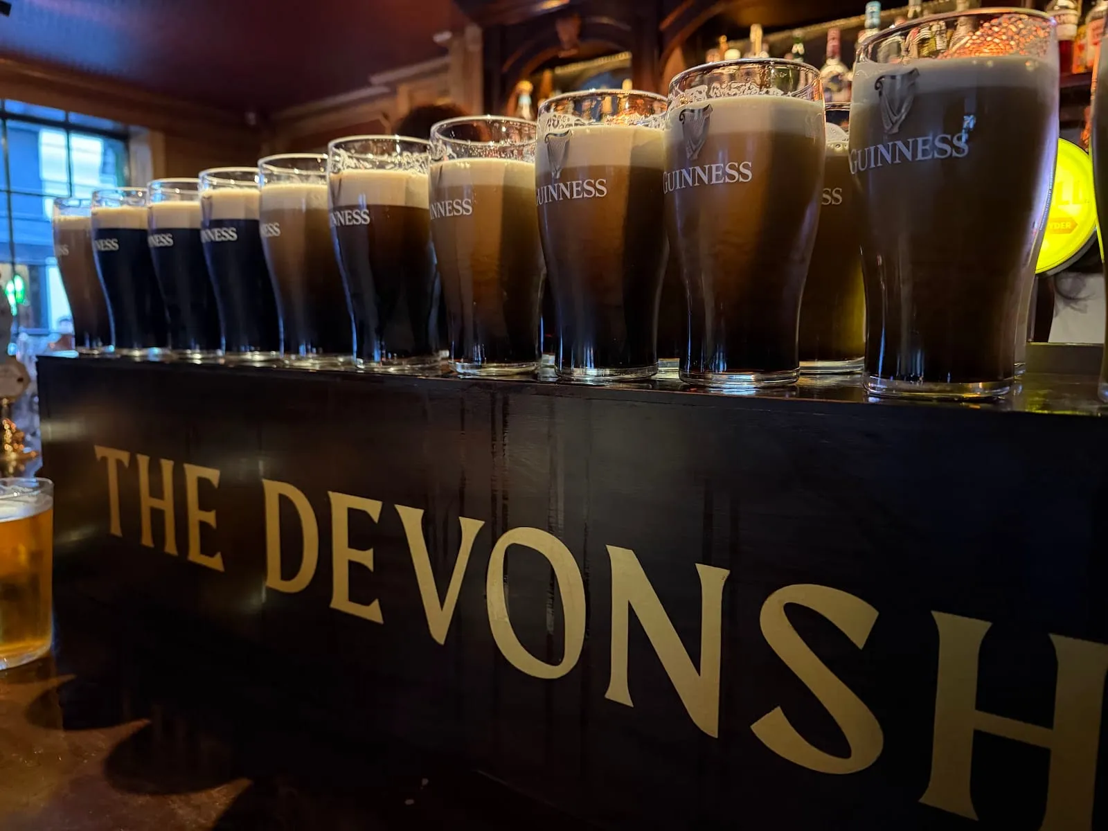A line of part-poured pints of Guinness settling along the bar top at The Devonshire, the pub's name in gold lettering across the bar front