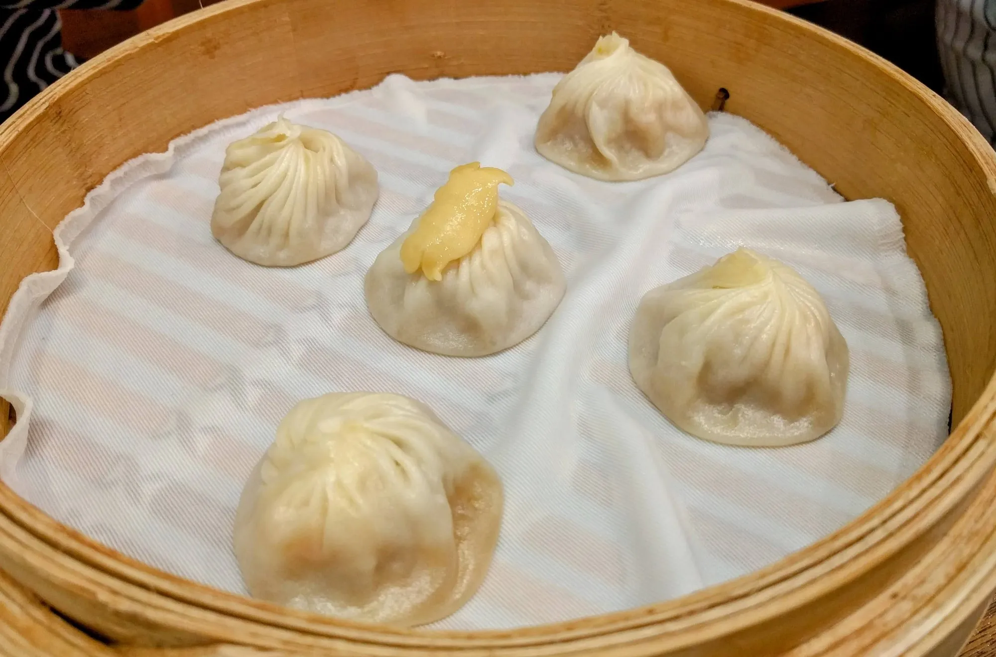 Five xiao long bao in a bamboo steamer lined with cloth, one topped with a spoonful of yellow crab roe