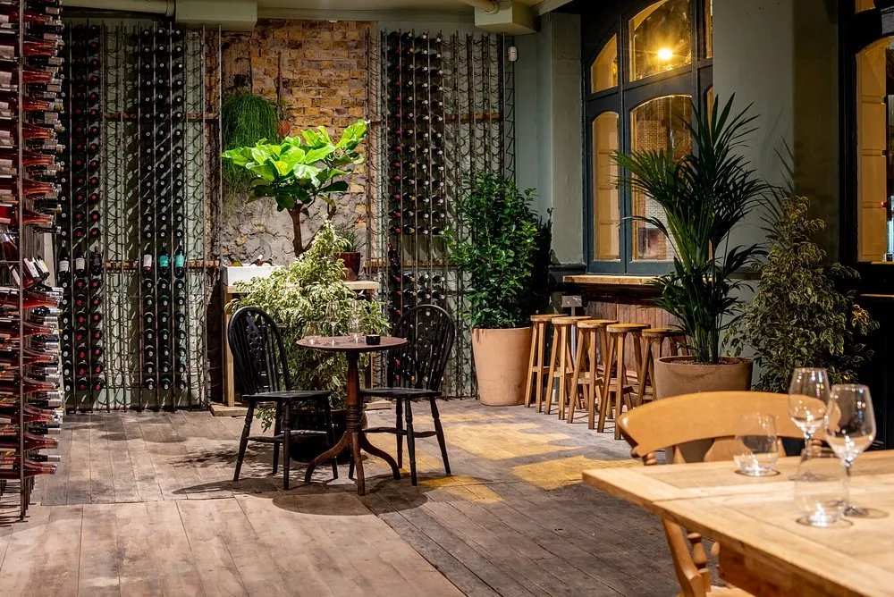 Floor-to-ceiling wine racks packed with bottles along a bare brick wall at Diogenes the Dog, with a small round table and bar stools on a wooden floor