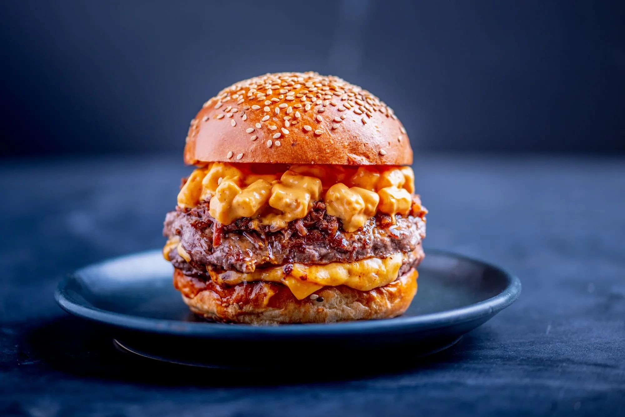 A double cheeseburger dripping with a chunky orange cheese sauce over a dark-seared patty, on a sesame seed bun on a dark plate