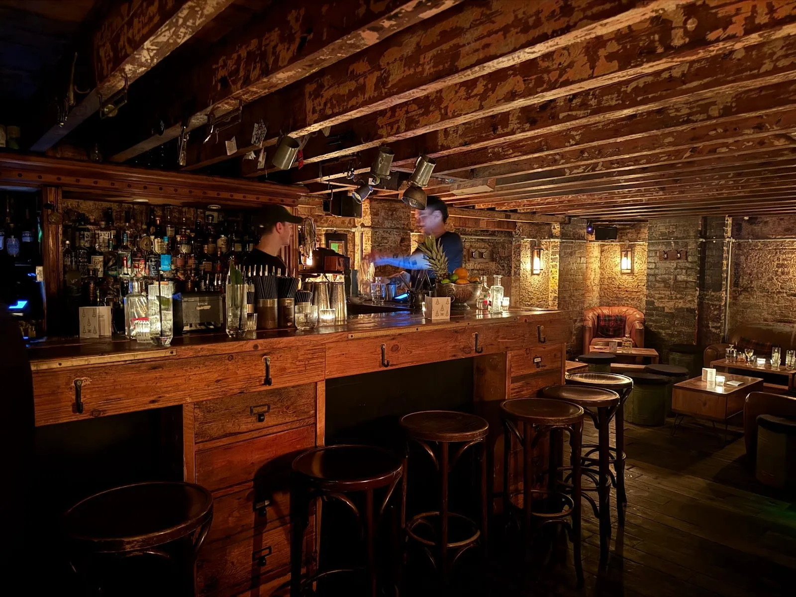 A long wooden bar with drawer fronts and iron hooks, bentwood stools, bare brick walls and old ceiling joists, with leather armchairs at the back of the room
