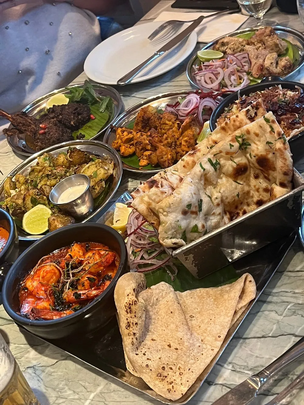 A crowded table at Dishoom holding lamb chops, chicken tikka, malai chicken, gunpowder potatoes, a prawn curry and a tin of naan on a marble top