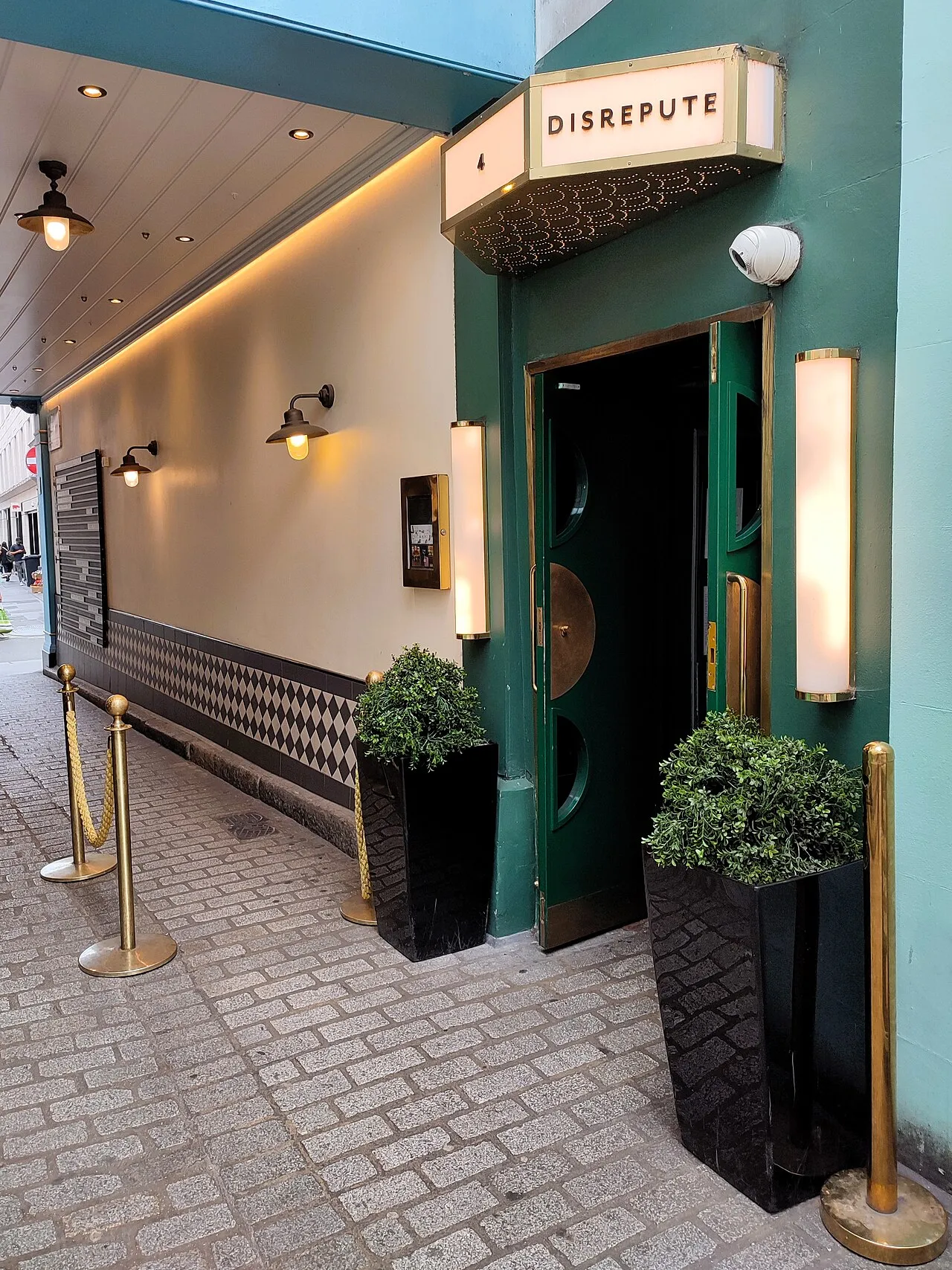 The green double doors of Disrepute at 4 Kingly Court, with DISREPUTE in a lit box sign above them, potted topiary either side and brass rope posts on the cobbles