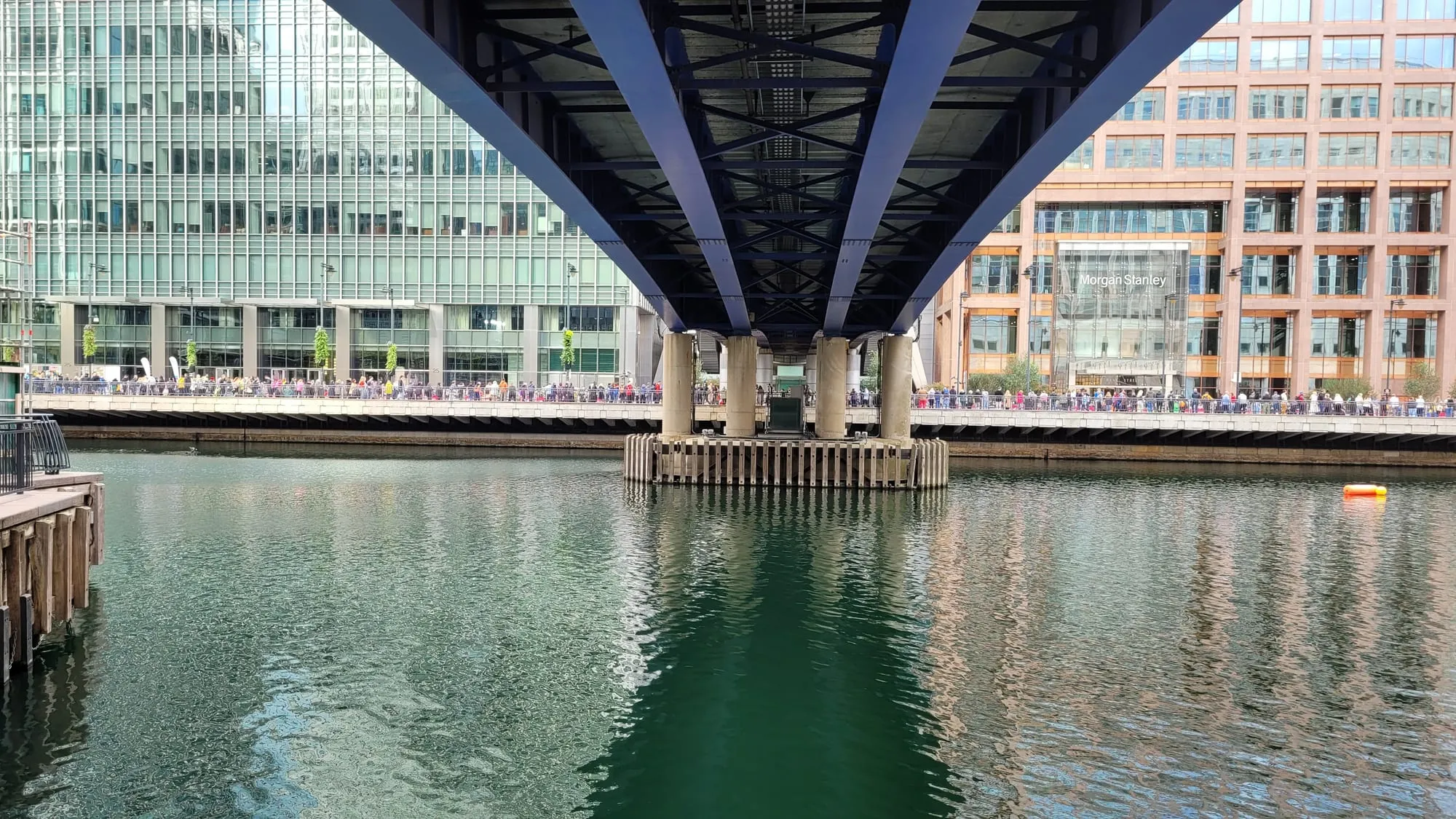 The DLR viaduct running between glass office towers over a Canary Wharf dock, with a crowd gathered below