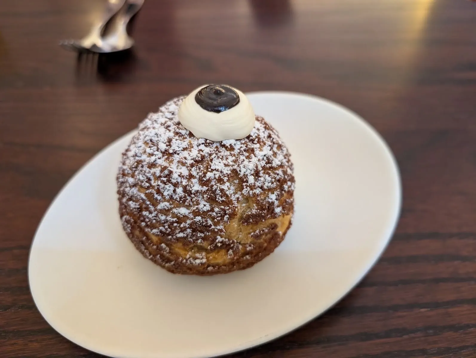 A round choux bun dusted heavily with icing sugar and topped with a piped white cream rosette and a dark glossy disc, on a white plate