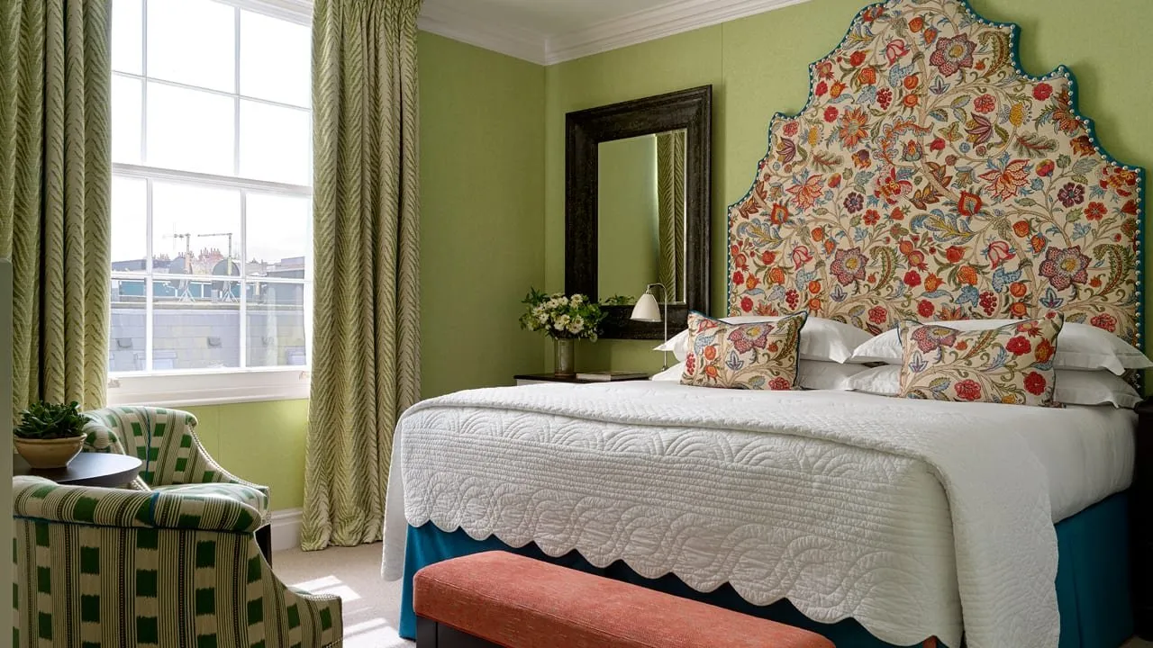 A bedroom at Dorset Square Hotel with green walls, a tall arched headboard embroidered with red and blue flowers, a scalloped white quilt, a green-striped armchair and a sash window over the rooftops