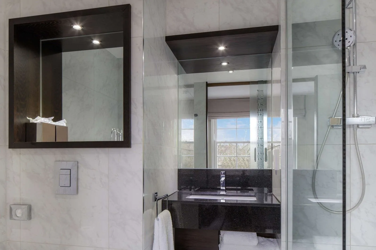 The bathroom at DoubleTree by Hilton London Greenwich: white marble-effect tiles, a black granite basin, a dark wood-framed mirror niche and a glass-walled shower printed with a timeline of Greenwich history