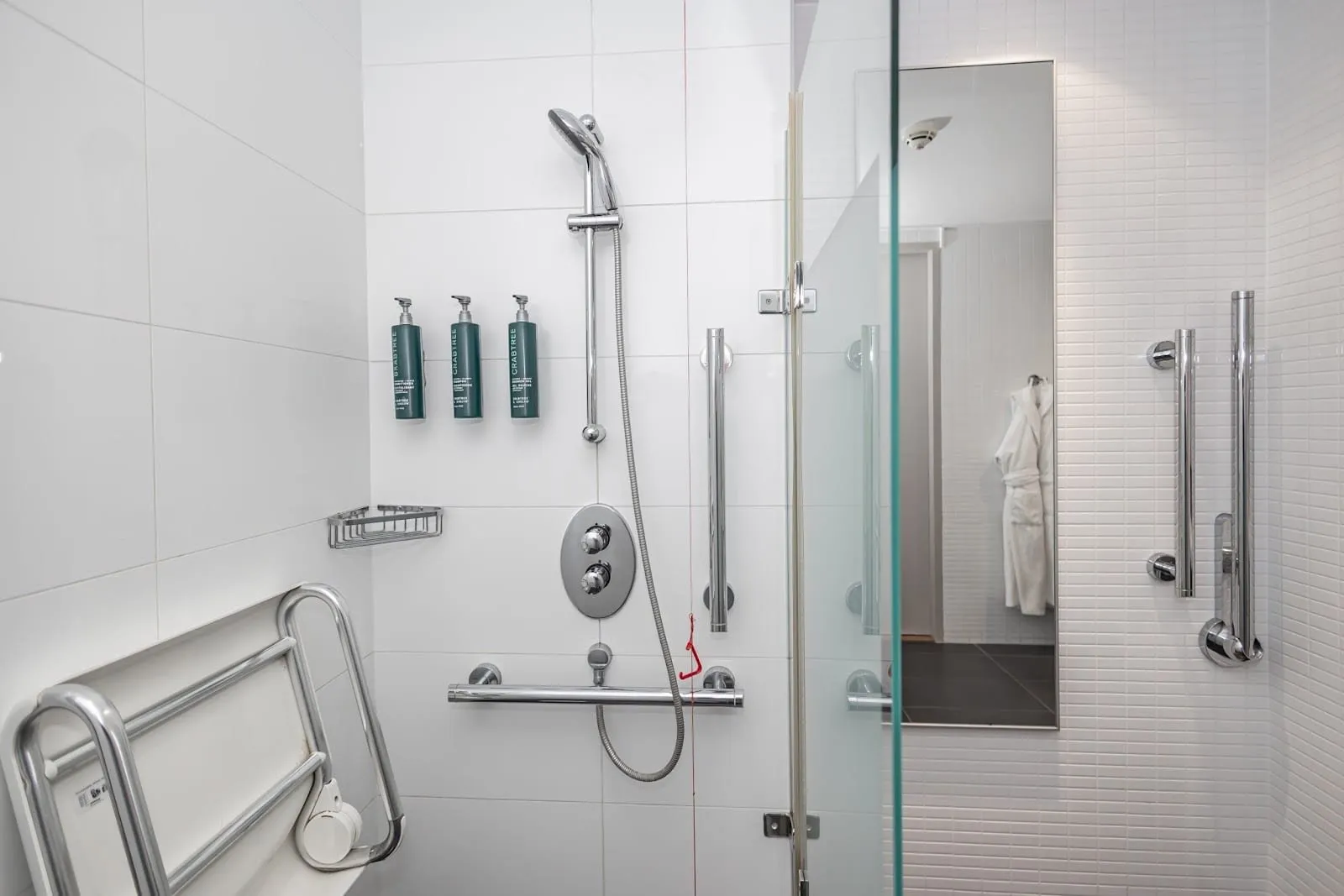 An accessible shower at DoubleTree Tower of London in white tiles, with a fold-down seat, grab rails, a handheld shower, a red alarm cord and Crabtree & Evelyn dispensers