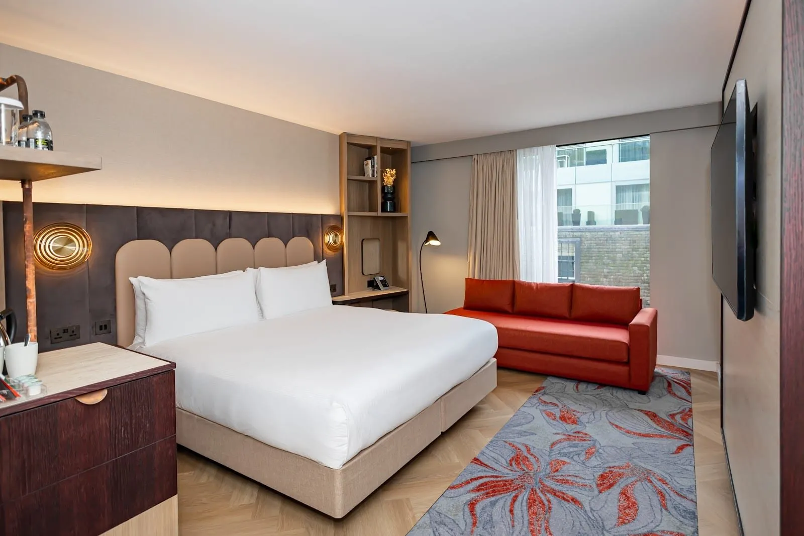 A king room at DoubleTree Tower of London with a scalloped beige headboard, brass wall lights, a red sofa by the window, a grey and red floral rug and herringbone floors