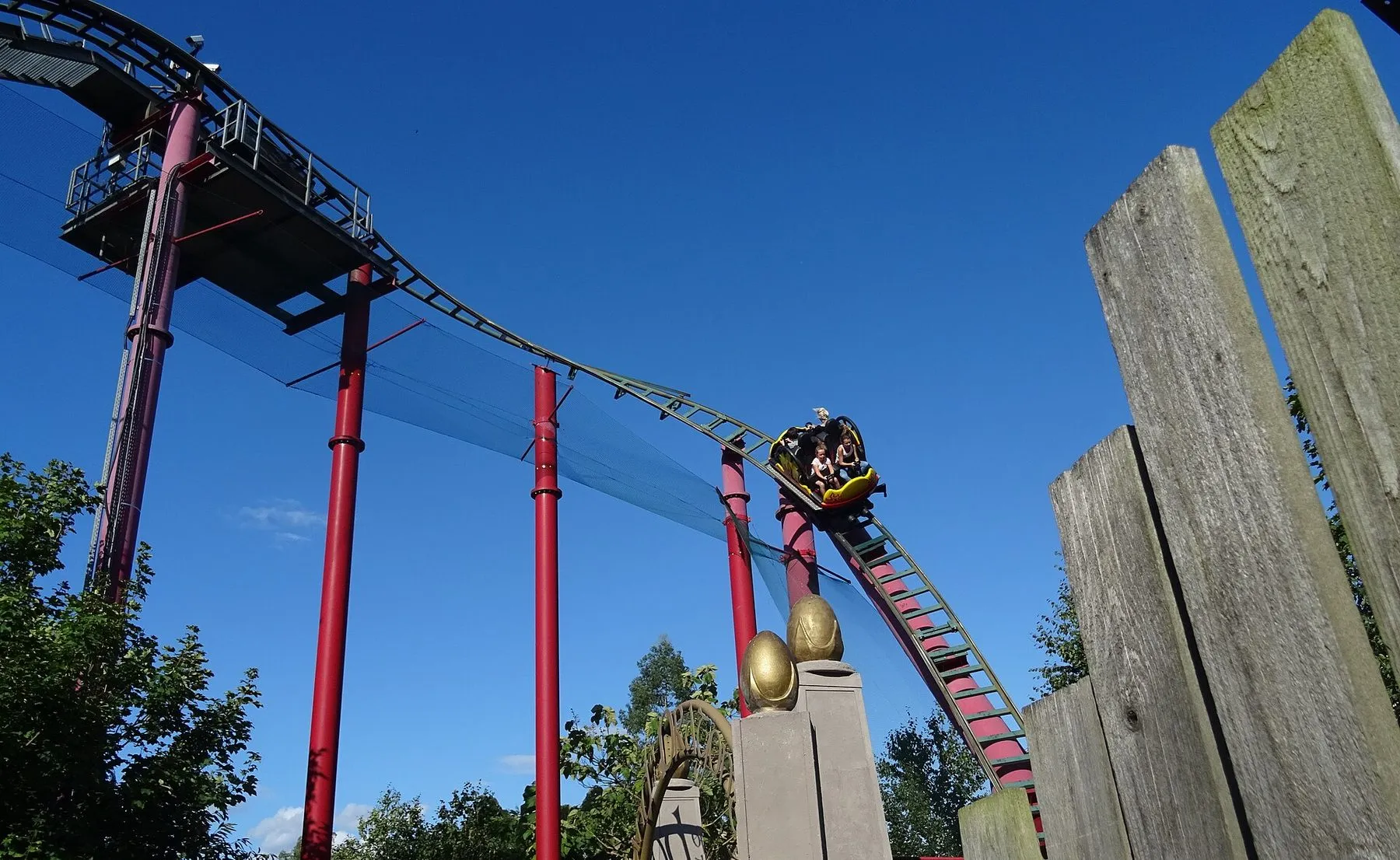 Dragon's Fury at Chessington, a car of riders spinning along red-supported track against a deep blue sky, golden dragon eggs on the gateposts below