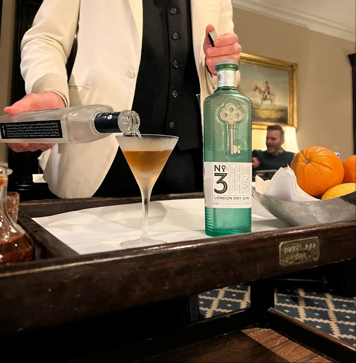 A bartender in a white jacket pouring from a frosted bottle into a martini glass on a wooden drinks trolley, beside a bottle of No.3 London Dry Gin and a bowl of citrus fruit