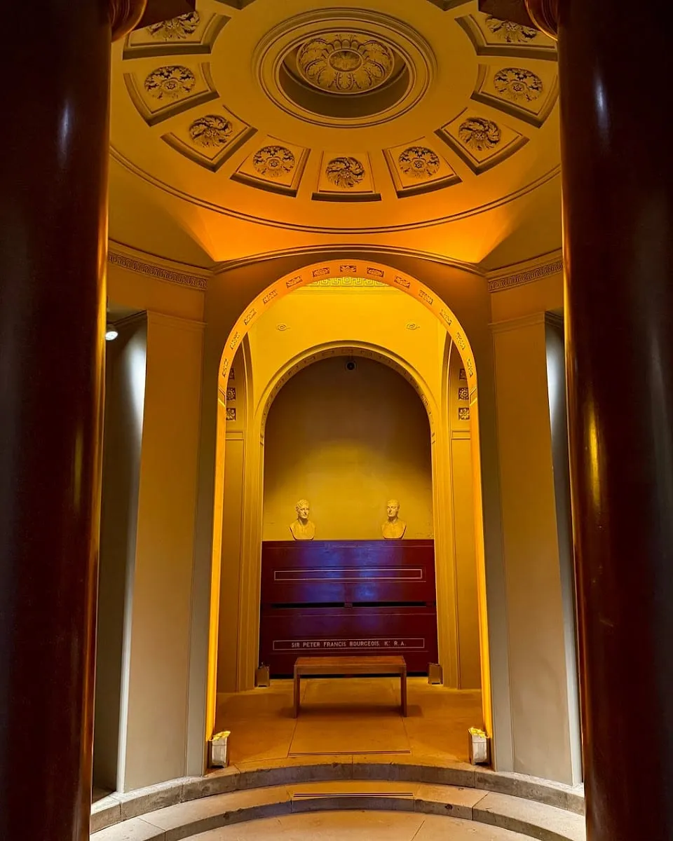 A domed mausoleum inside Dulwich Picture Gallery, with two marble busts on a dark sarcophagus lit in gold light