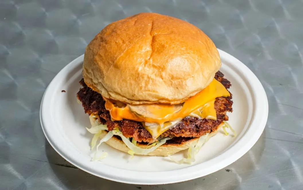 A smash burger on a white paper plate on a steel counter, two crisp-edged patties under melted American cheese and sauce, with shredded lettuce showing at the base of the bun