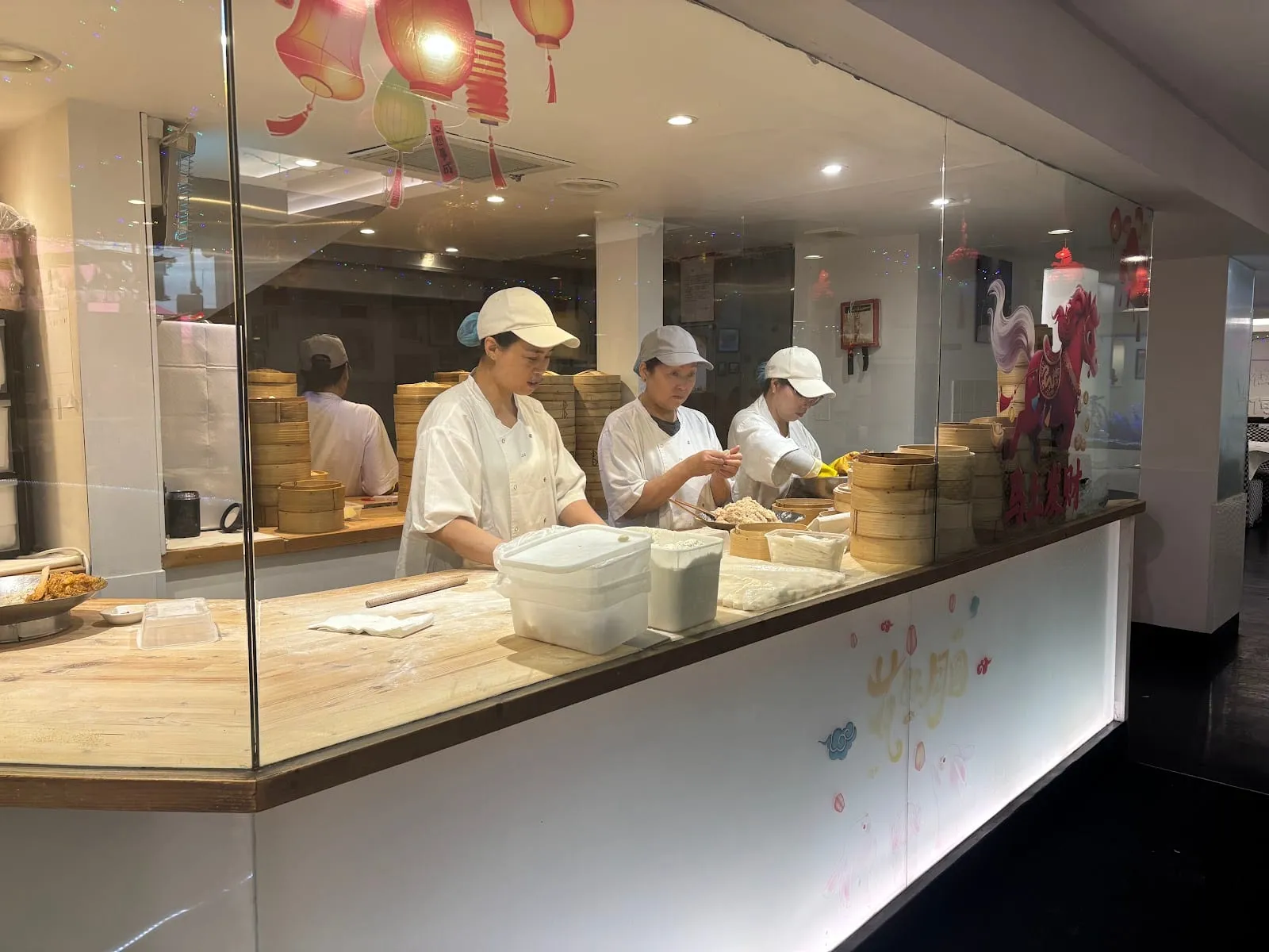 Staff in white uniforms and caps folding dumplings behind a glass-walled kitchen counter, with stacks of bamboo steamers
