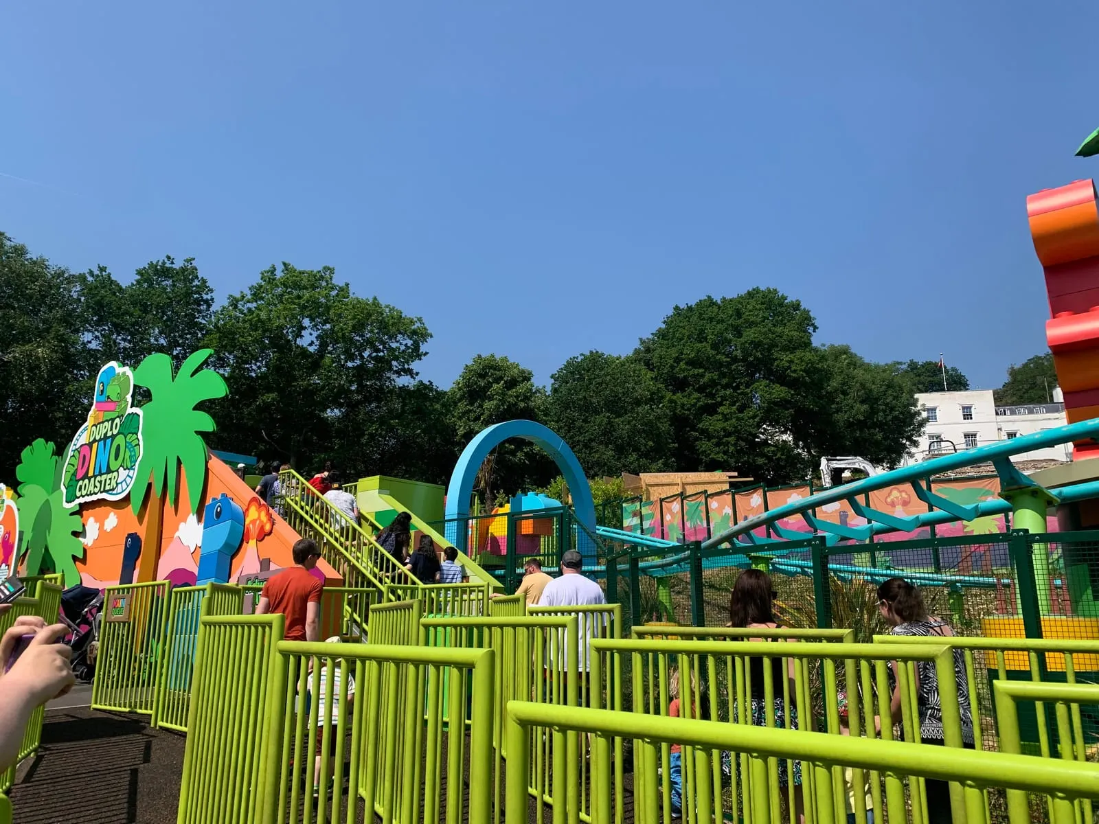 The DUPLO Dino Coaster in DUPLO Valley, its bright green and orange queue line and track among palm-shaped decorations under a blue sky