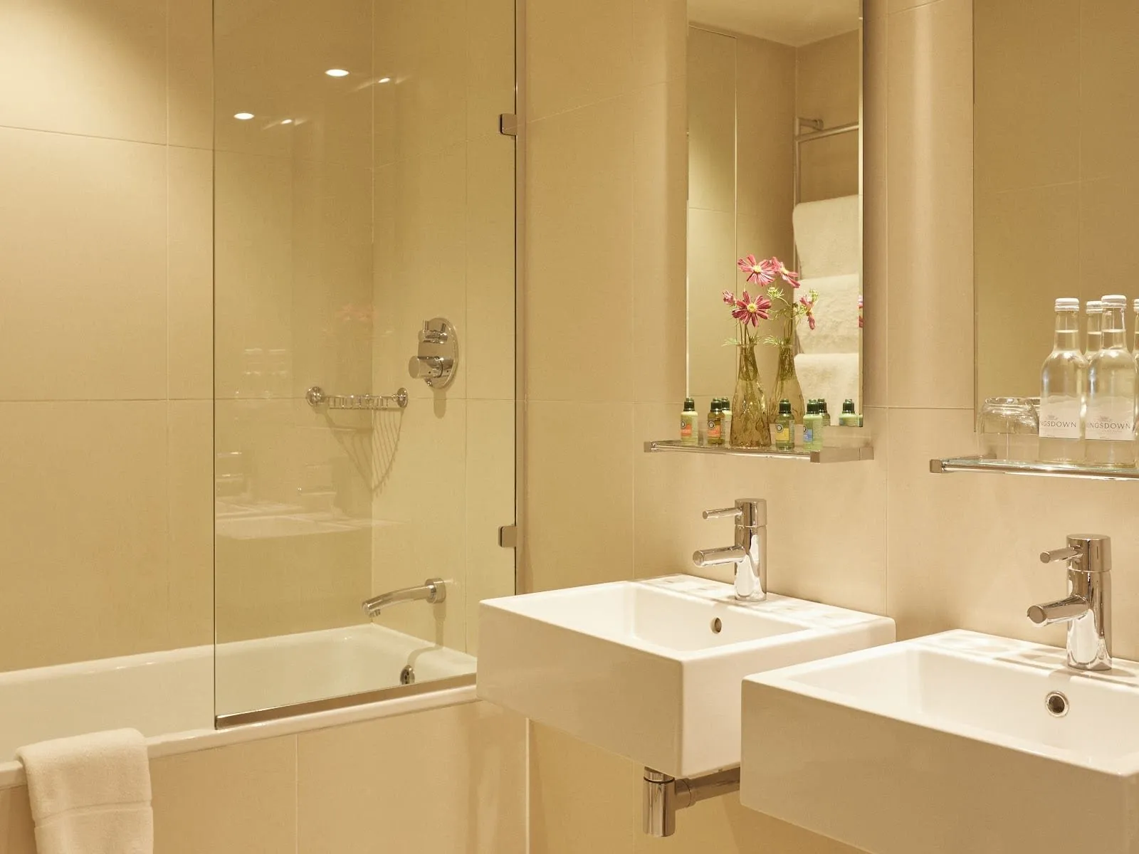 A cream-tiled bathroom at Durrant's with a bath behind a hinged glass shower screen and two square white basins side by side