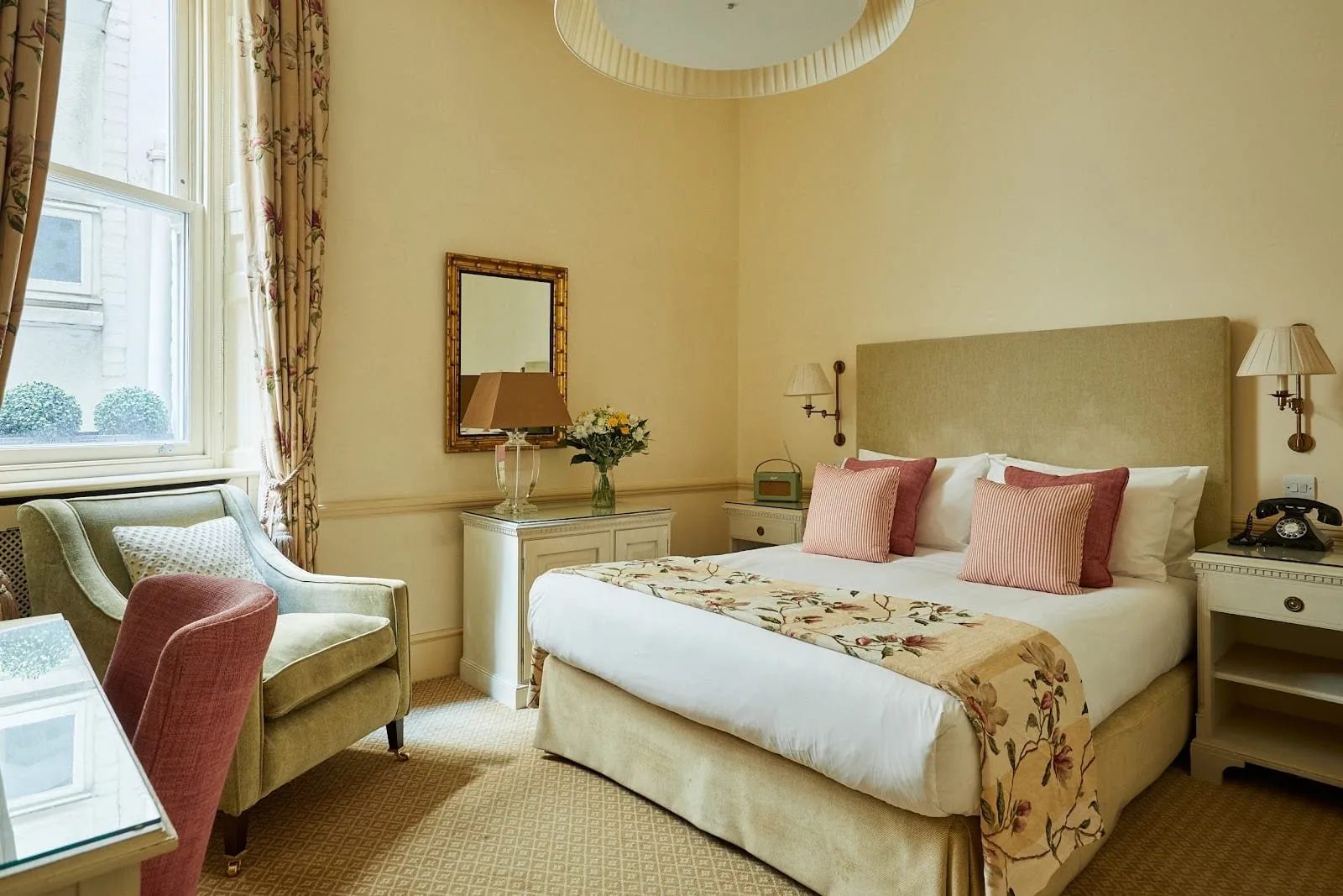 A double room at Durrant's with cream walls, a green upholstered headboard, a floral bed runner, a sage armchair by a tall sash window and a black dial telephone on the bedside table