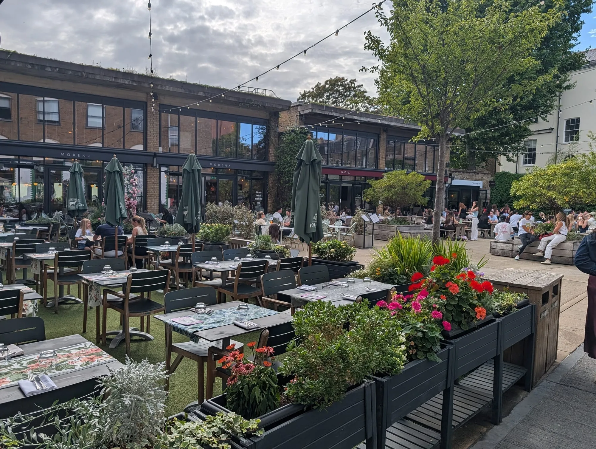 Outdoor tables and planters in the courtyard at Eccleston Yards, with brick warehouse buildings behind