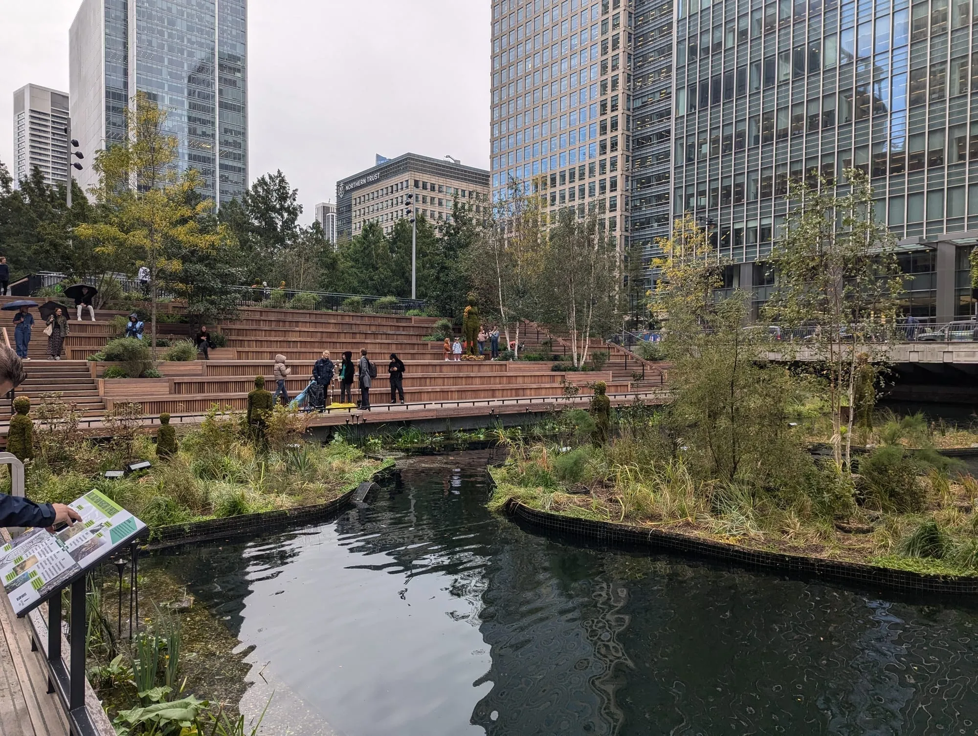 Floating planted islands and wetland walkways at Eden Dock, with Canary Wharf towers rising behind