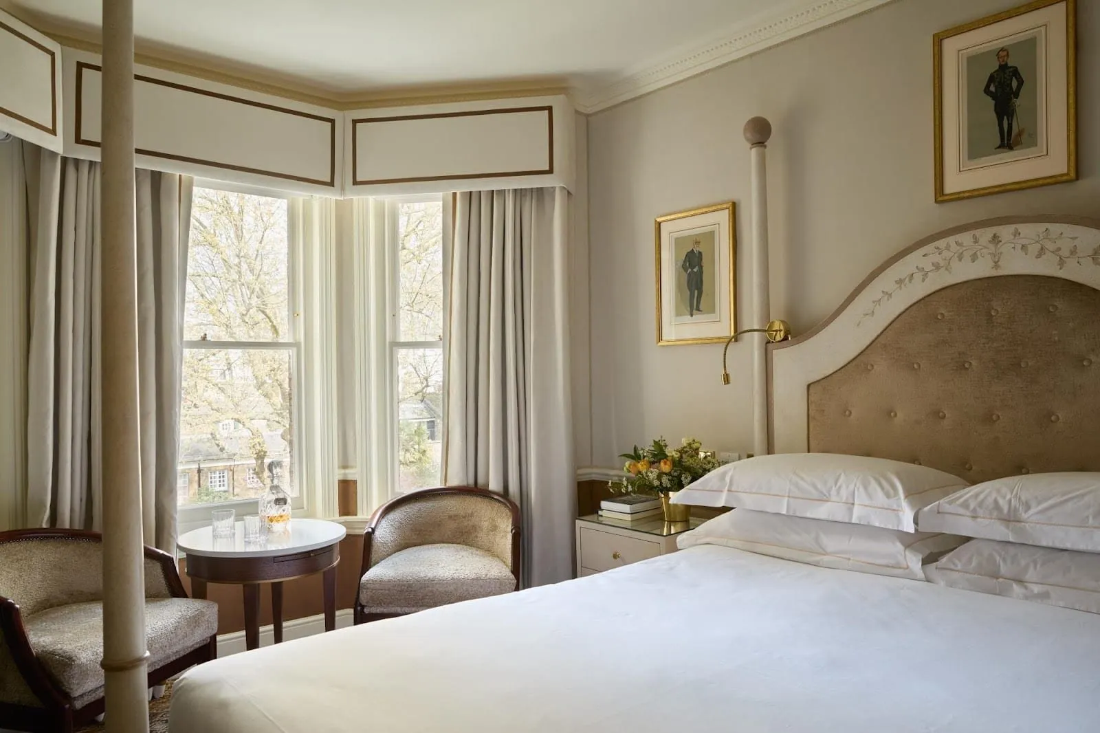A Deluxe Queen room at Egerton House: a four-poster bed with a buttoned velvet headboard, framed caricature prints, two tub chairs at a round table and a bay of sash windows onto bare trees