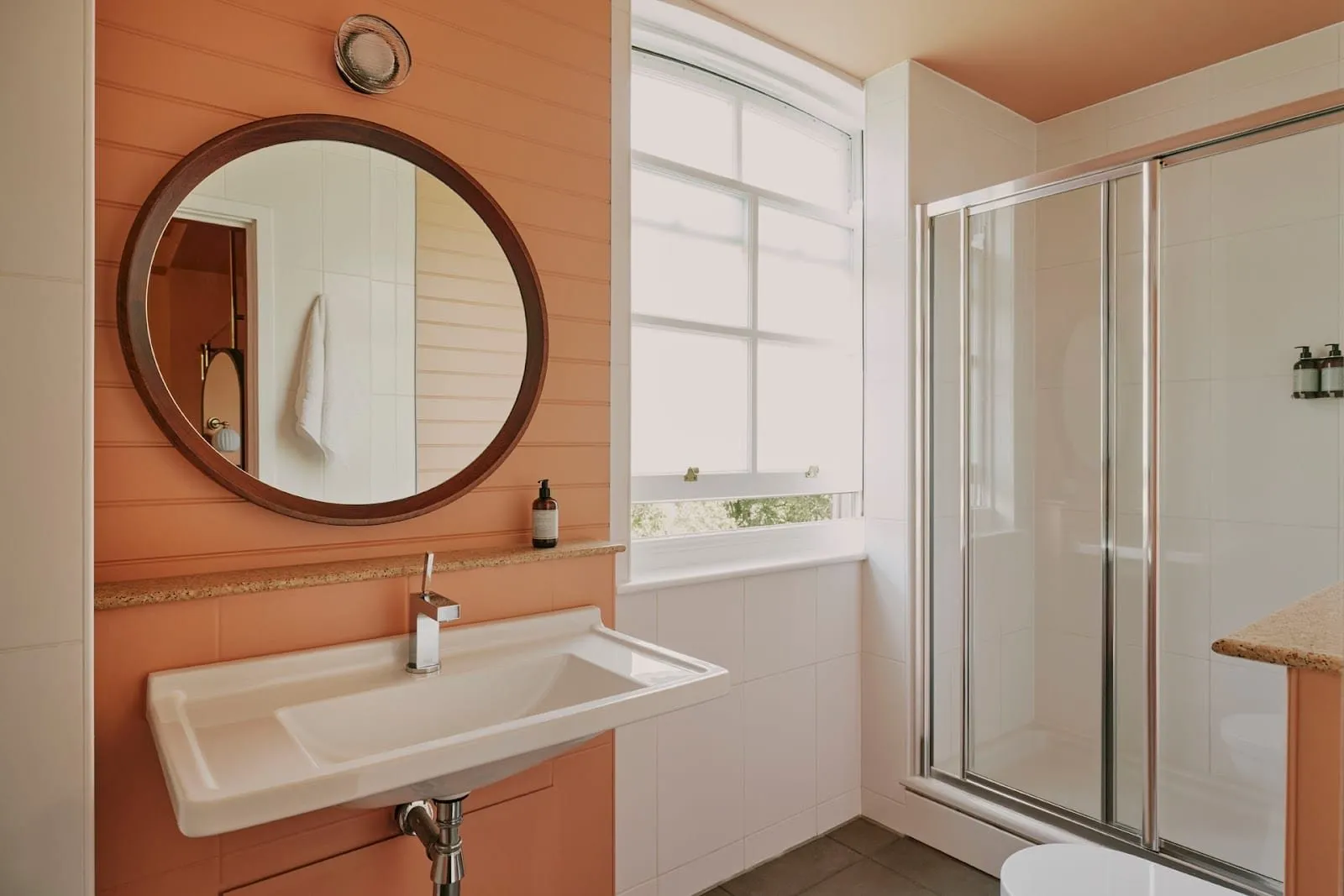 An Ember Locke shower room with terracotta tongue-and-groove panelling, a round wood-framed mirror over a wide basin, a sash window and a glass-doored shower