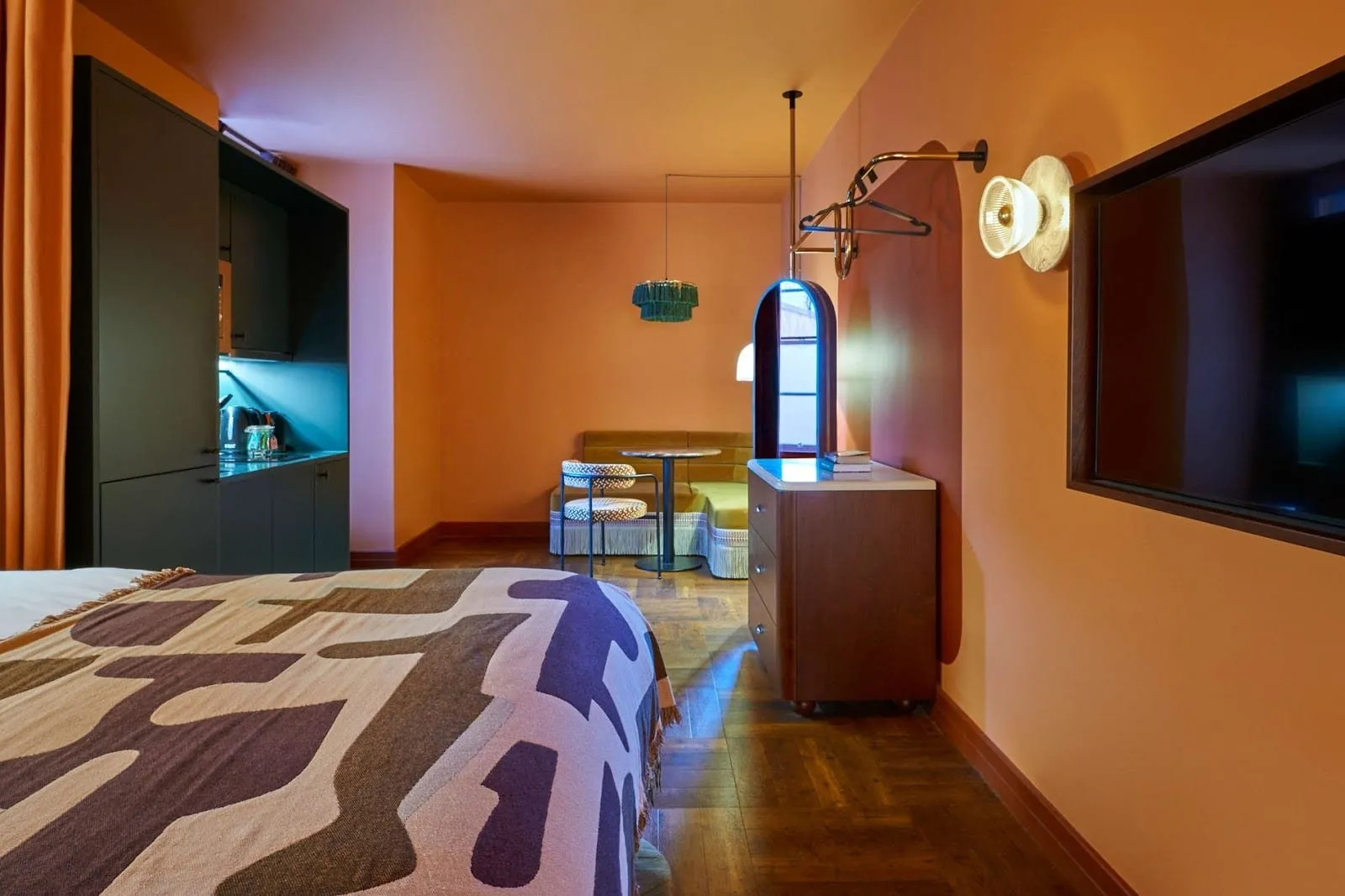 An Ember Locke studio with terracotta walls, a dark green kitchenette with a sink, kettle and built-in microwave, a fringed banquette dining nook, a walnut chest of drawers and an arched mirror, seen from beside the bed