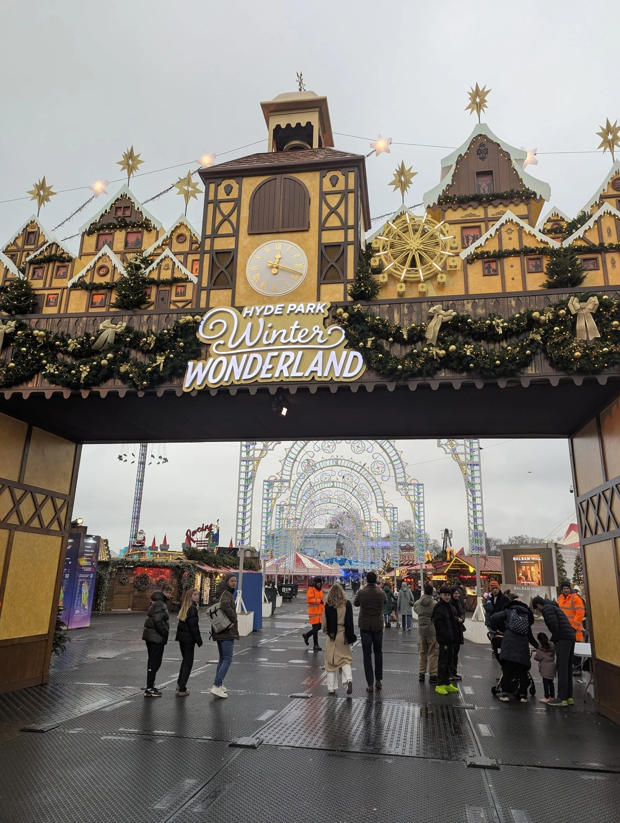 The Bavarian-style entrance gate to Hyde Park Winter Wonderland, with a working clock tower and illuminated stars above the sign, and the lit arches of Luminarie Lane visible through the arch