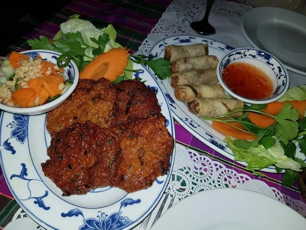 Fried fishcakes on blue-and-white patterned china beside a plate of spring rolls with sweet chilli dipping sauce, a cucumber and peanut relish and a carved carrot garnish, on a patterned tablecloth