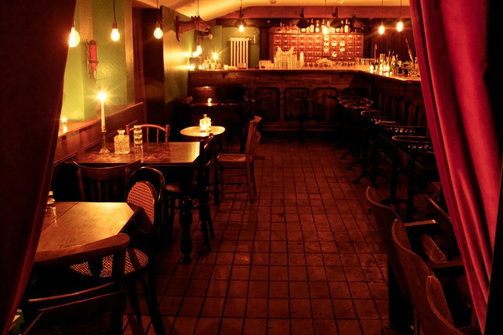 The basement bar at Evans & Peel seen past a red velvet curtain, with candlelit tables, stools along a wooden counter and a tiled floor