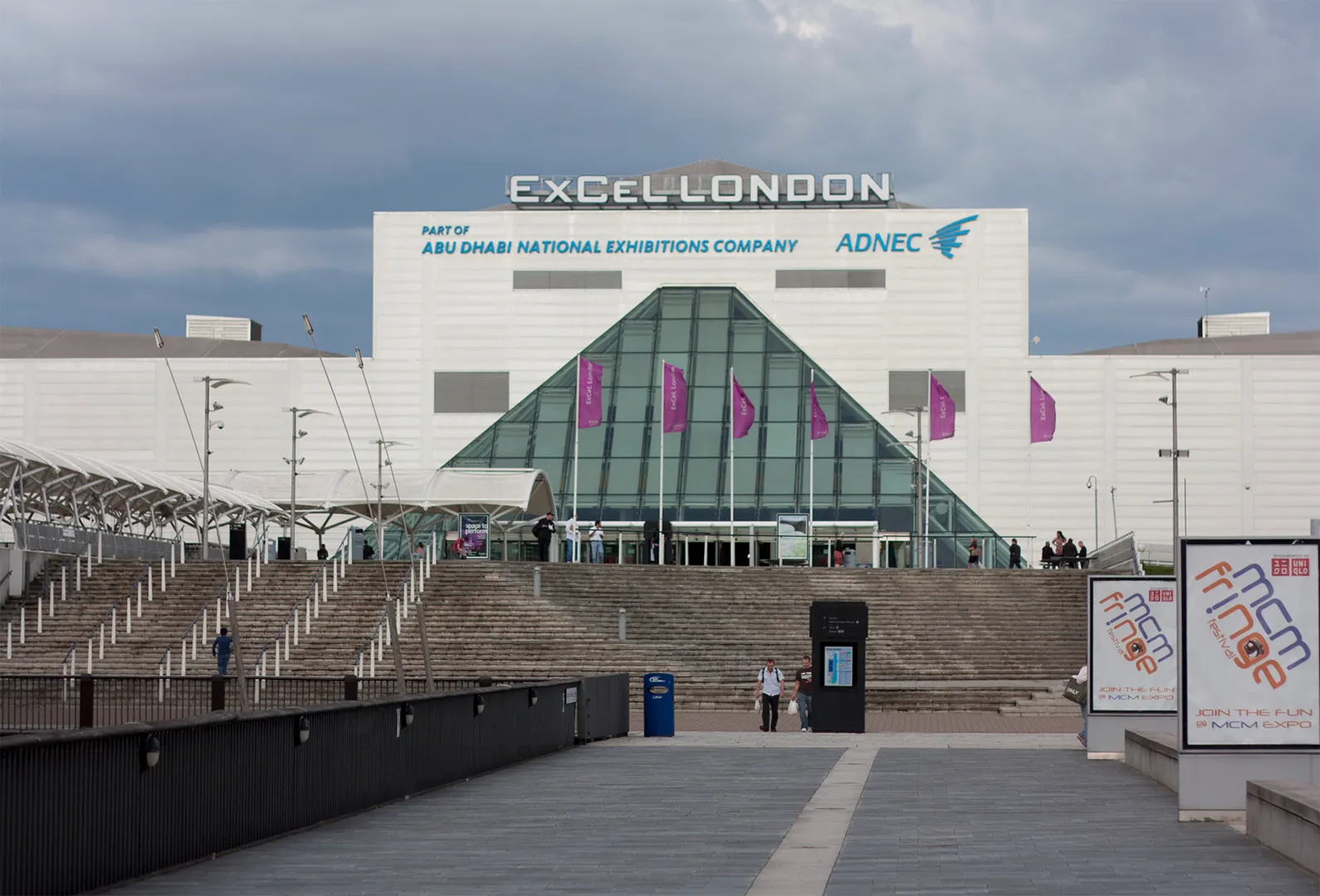 ExCeL London's glass entrance pyramid and rooftop sign, seen across the plaza with event flags flying