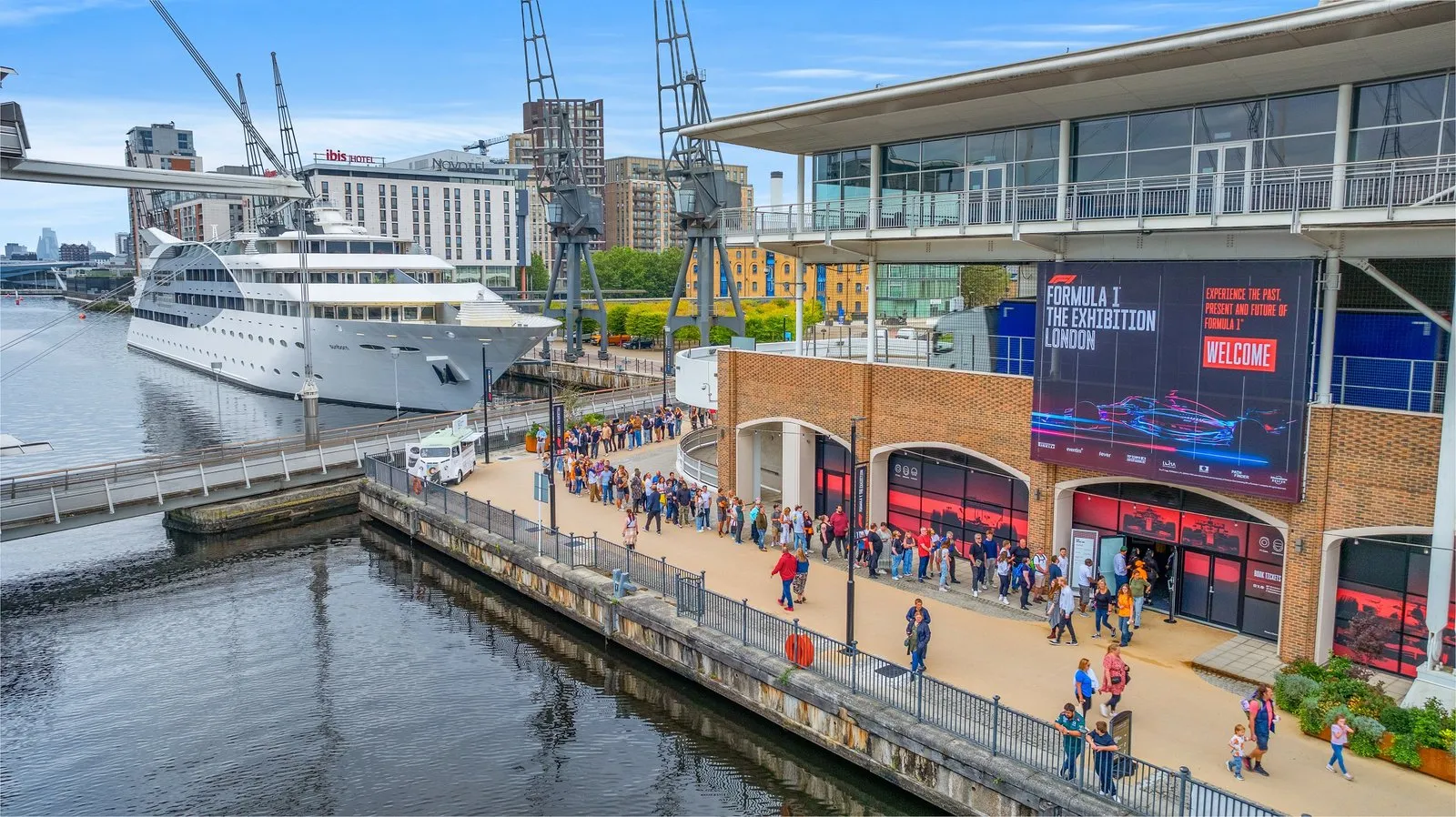 Visitors queuing beside the Royal Victoria Dock outside ExCeL London, with dockside cranes, moored boats and a large screen advertising a show at the entrance