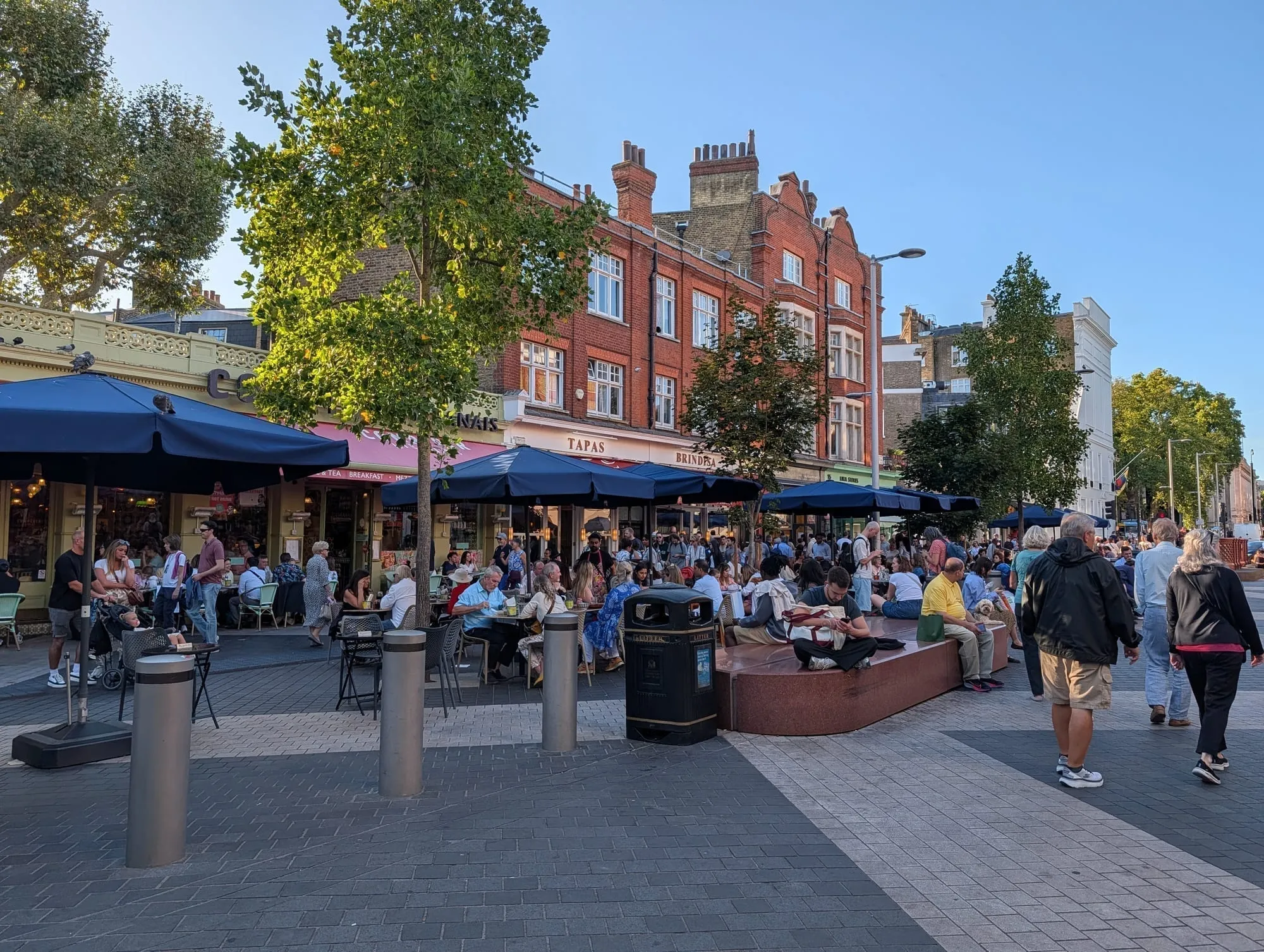 Diners at outdoor tables along Exhibition Road in the evening, with red brick buildings behind
