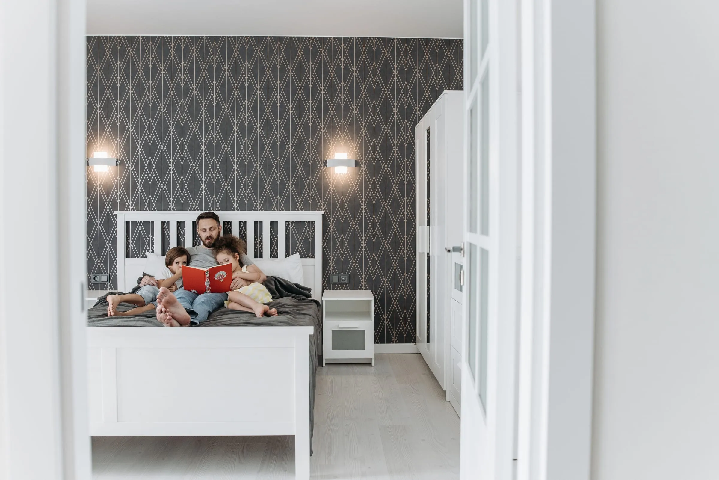 A father reading a red picture book to two young children on a double bed, seen through an open bedroom door
