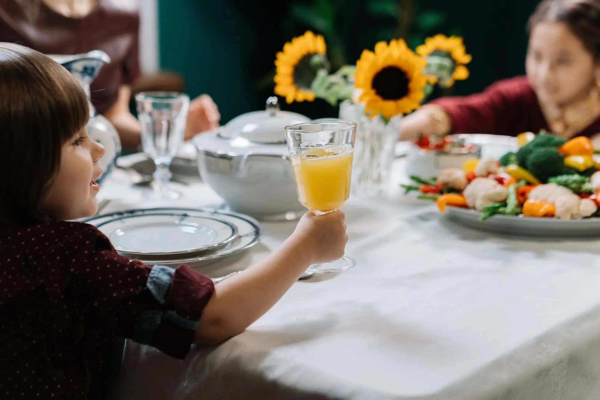 A young child at a laid dining table raising a glass of orange juice, with a platter of vegetables and family members around them