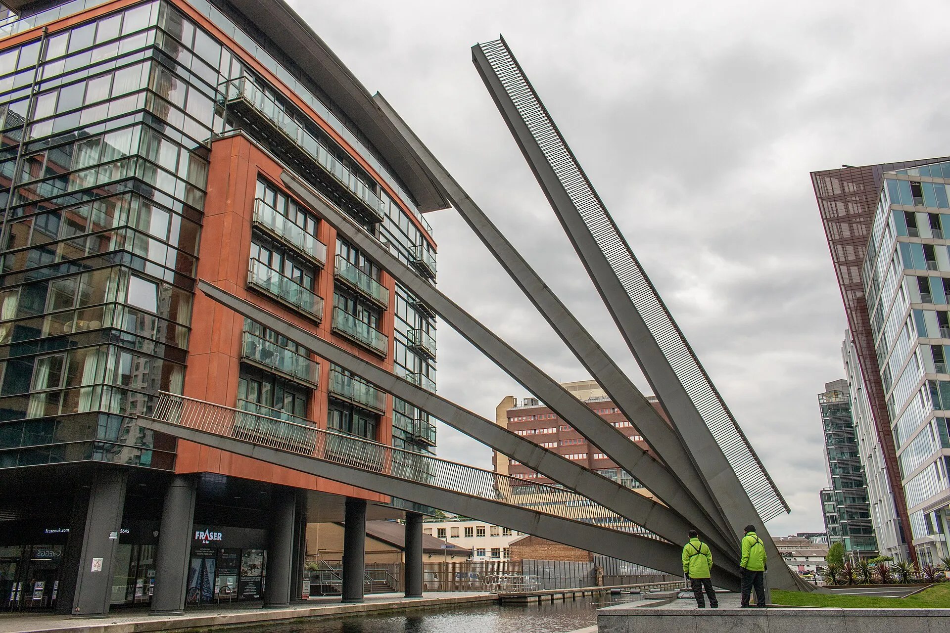 The Fan Bridge at Paddington Basin raised into its fan shape, steel fins angled up against a grey sky beside a red-brick apartment block