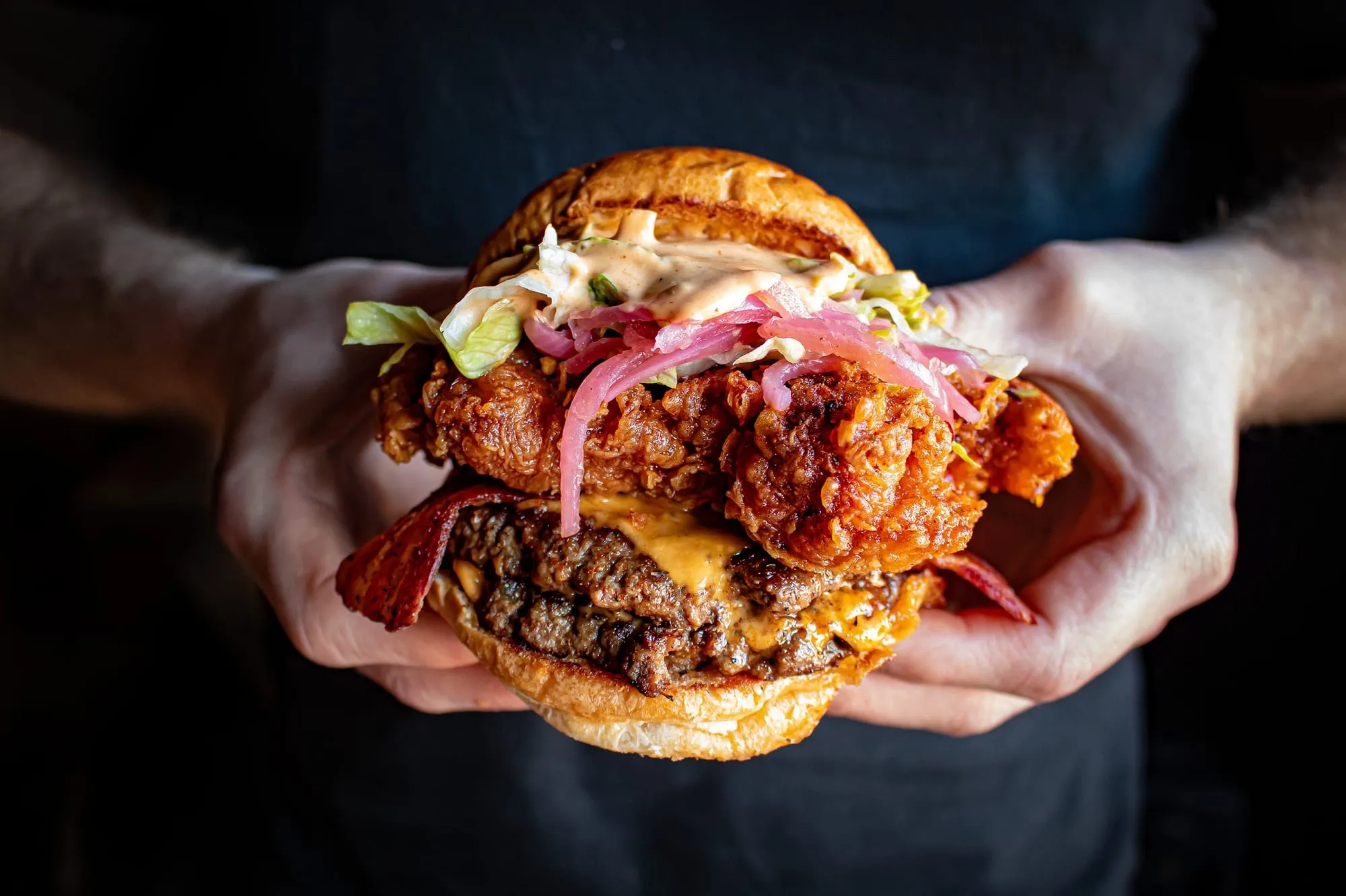 A tall burger held in two hands, with a dark-seared double beef patty, crispy fried chicken pieces, bacon, pickled onion and a pale sauce spilling from a toasted bun