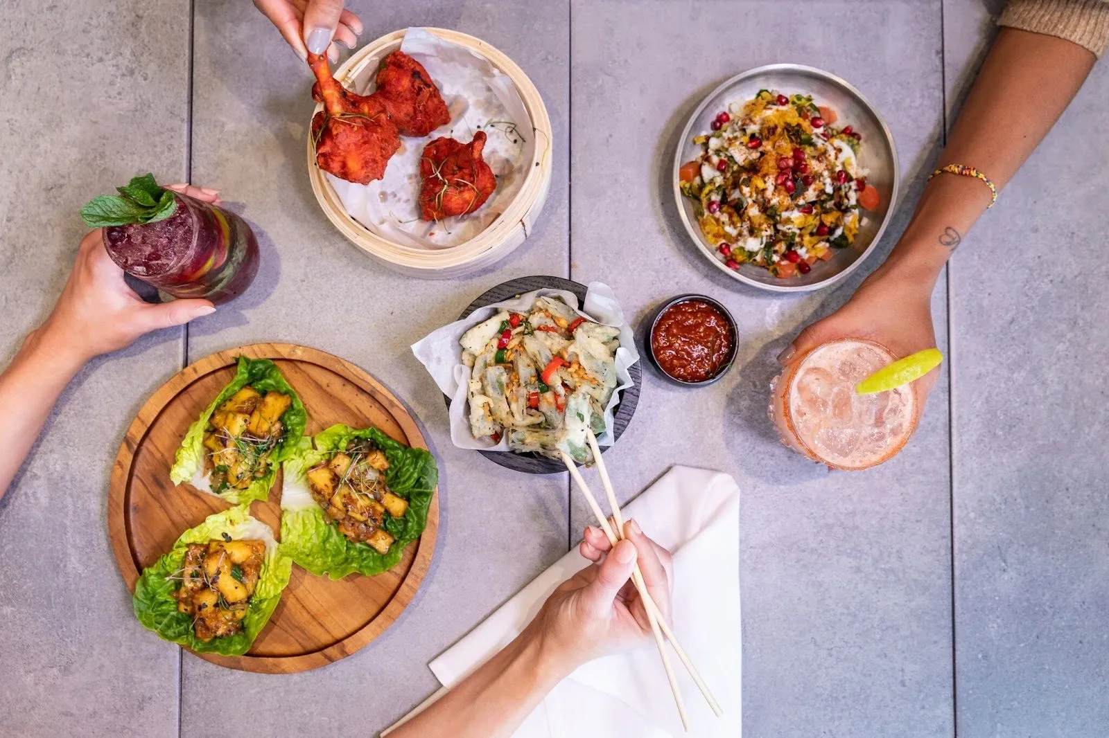 Fatt Pundit's small plates from above: chilli chicken lollipops in a bamboo steamer, paneer in lettuce cups, battered crackling spinach with chilli, a bowl of chaat with pomegranate, and two cocktails