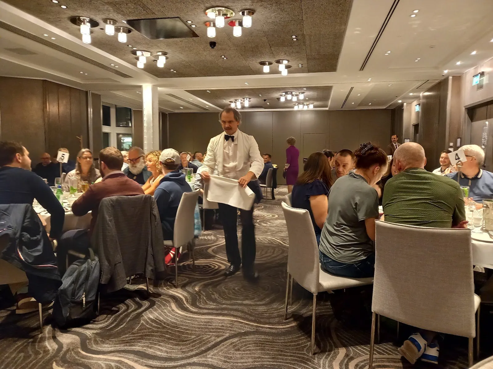 Manuel, in a white waiter's jacket, walks between diners at numbered tables in a hotel function room, with Sybil and Basil behind