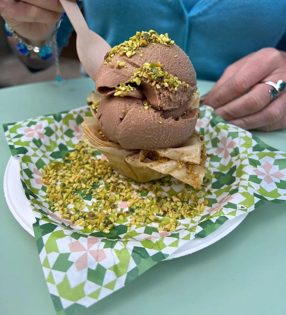 Two scoops of pistachio-flecked gelato served in a kunafa pastry cup, showered with crushed pistachios on patterned paper