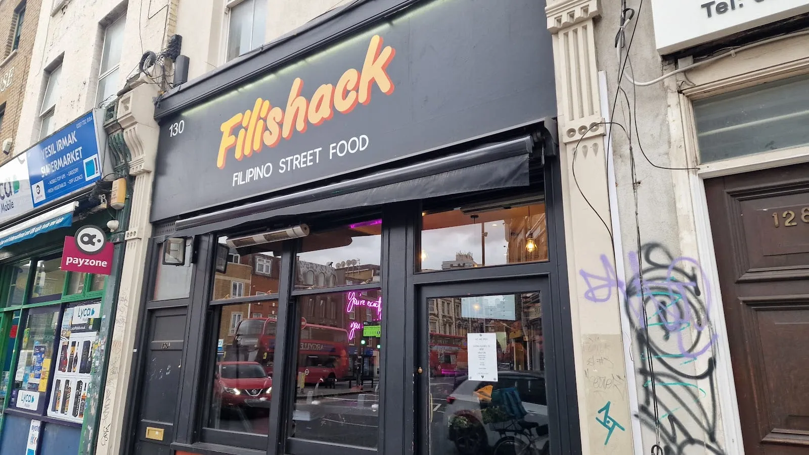 The Filishack shopfront at number 130: a dark fascia with the name in orange script above "Filipino street food", a plate-glass window with a pink neon sign inside, and a corner shop next door