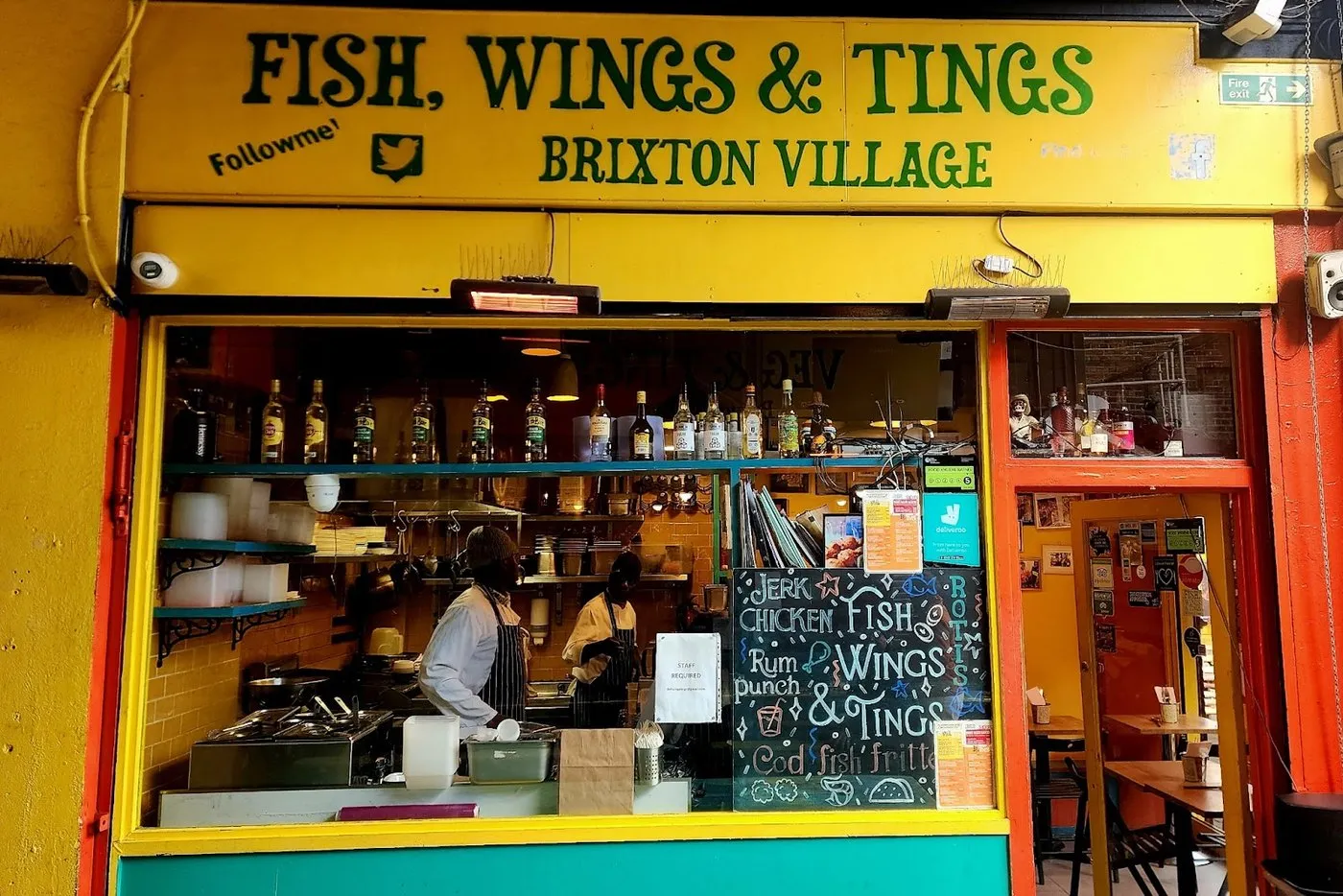The counter at Fish, Wings & Tings in Brixton Village, yellow and green signage above an open kitchen with two chefs working, and a chalkboard listing jerk chicken, rotis and cod fish fritters