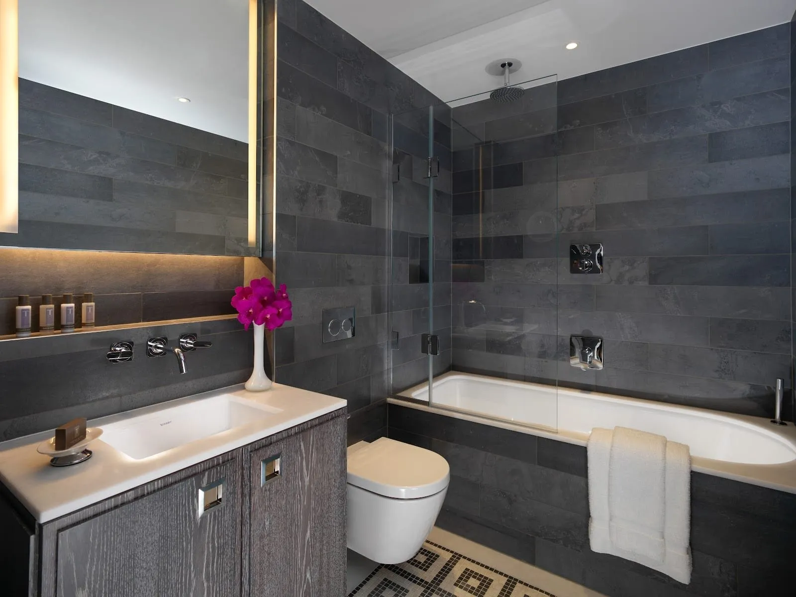 A Flemings Mayfair bathroom in grey slate tiles, with a bath under a glass screen and ceiling rainfall shower, a wall-hung WC and a basin on a grey oak vanity