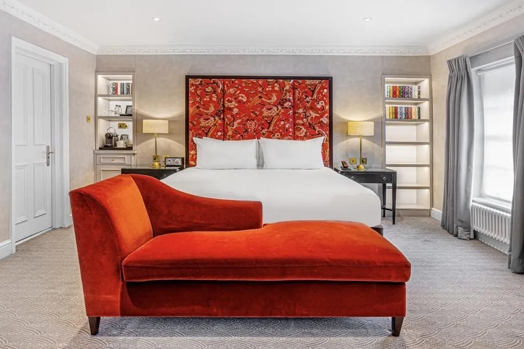 A Flemings Mayfair suite bedroom with a red bird-print headboard, a rust velvet chaise longue at the foot of the bed, lit bookshelves either side and a sash window above a white radiator