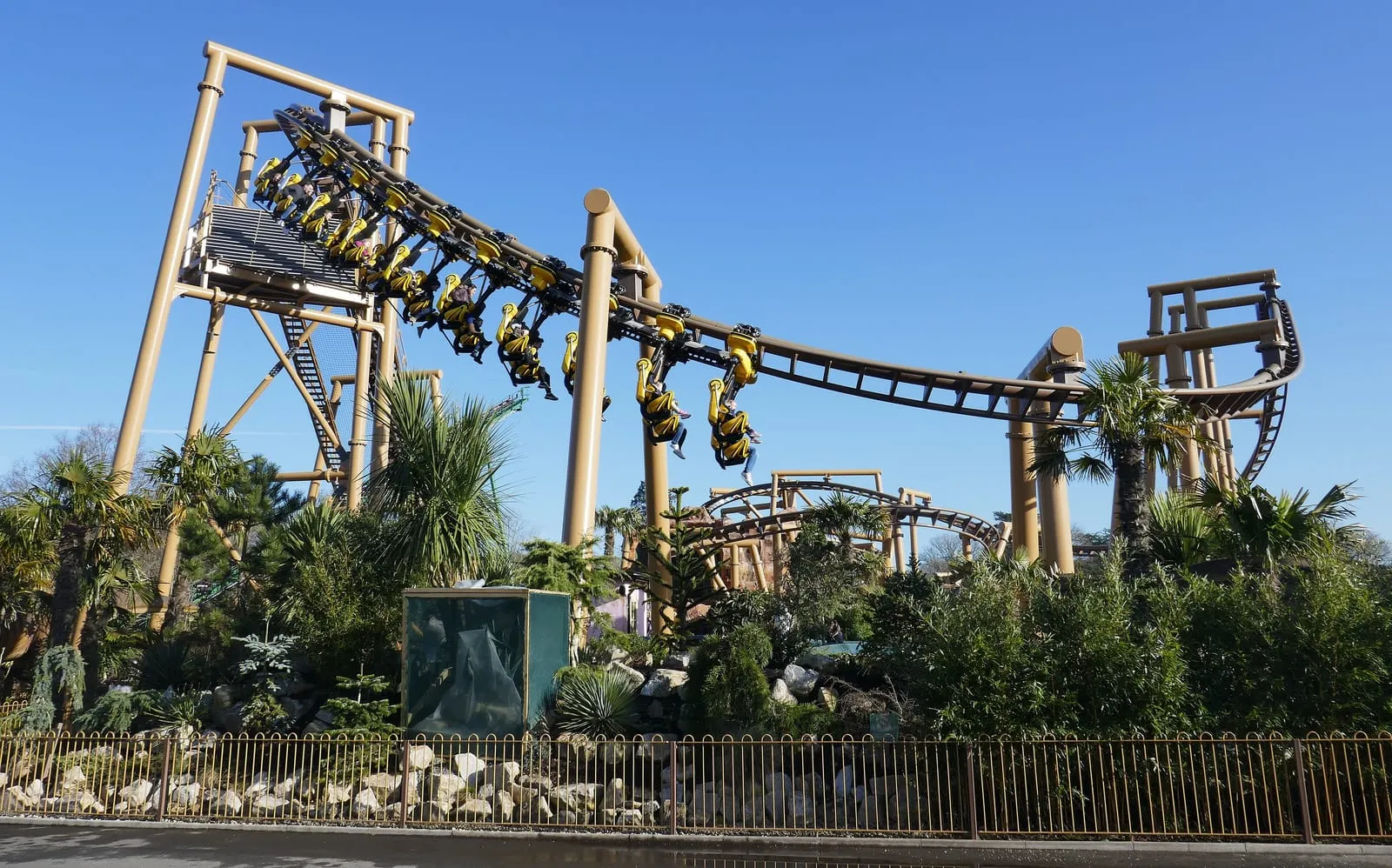 Flight of the Pterosaur at Paultons Park, riders hanging in yellow seats beneath the beige track as it banks above palm trees and rockwork